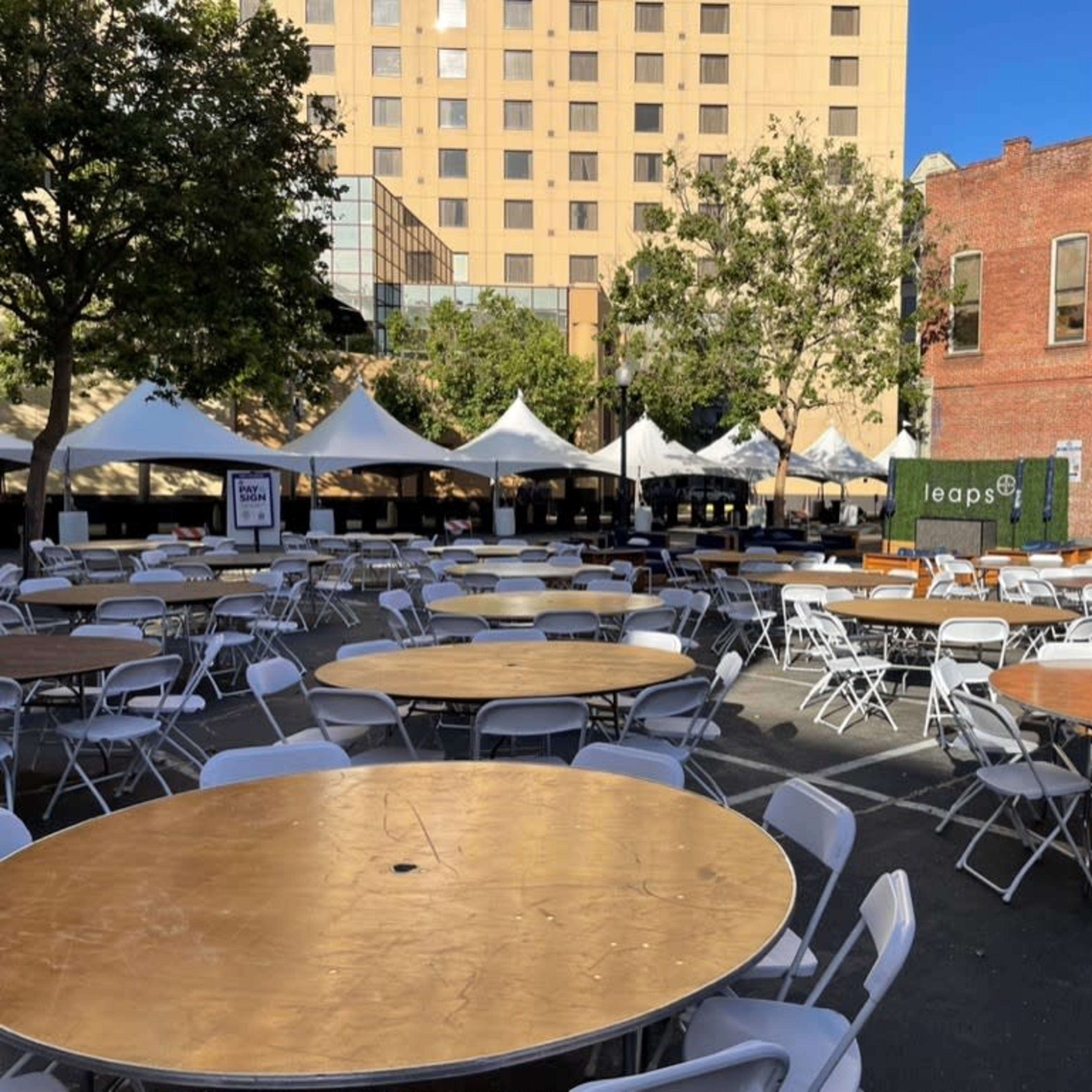 A spacious outdoor area filled with circular tables and white chairs, surrounded by tents and buildings.