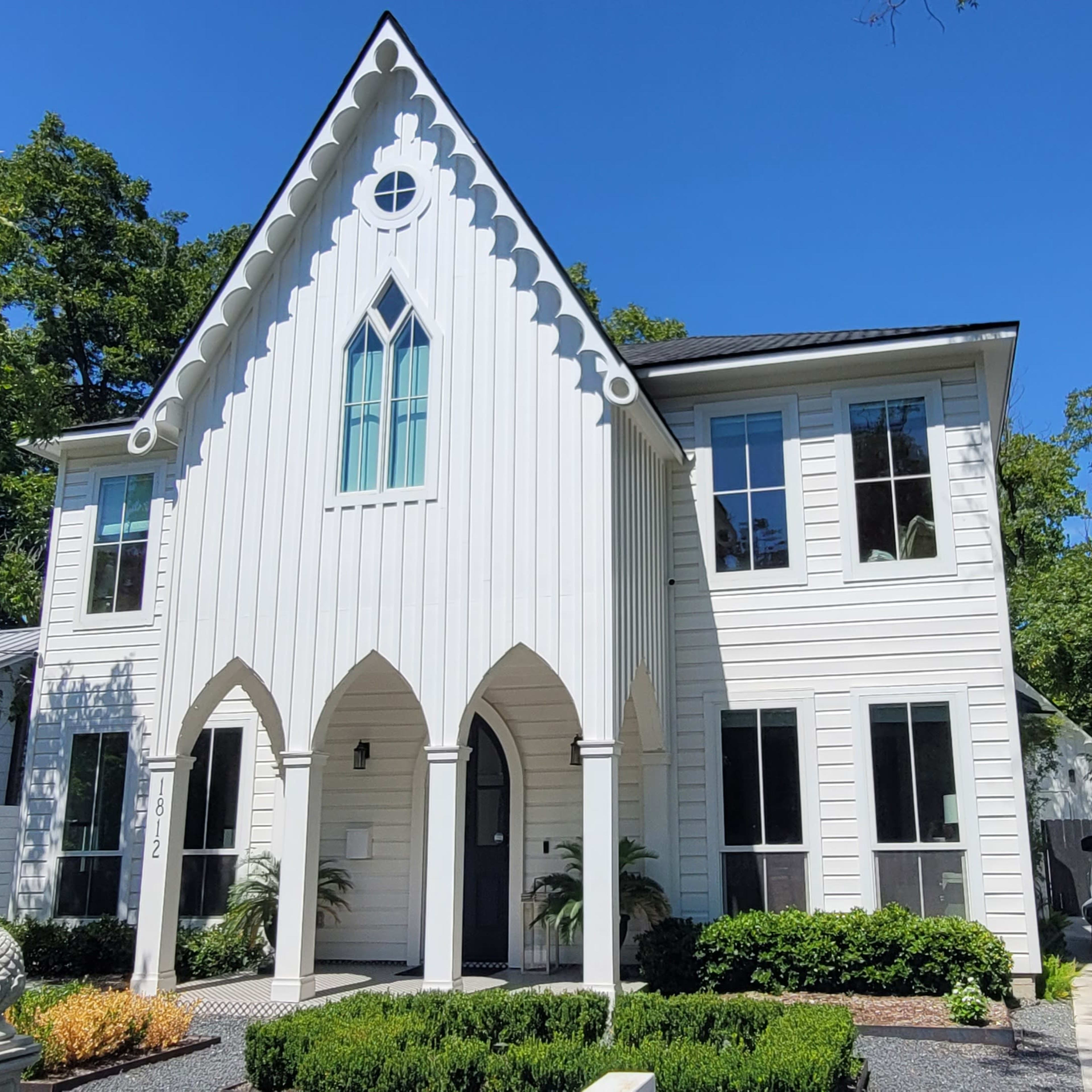 A white, Gothic-style house with a peaked roof and large windows surrounded by greenery.