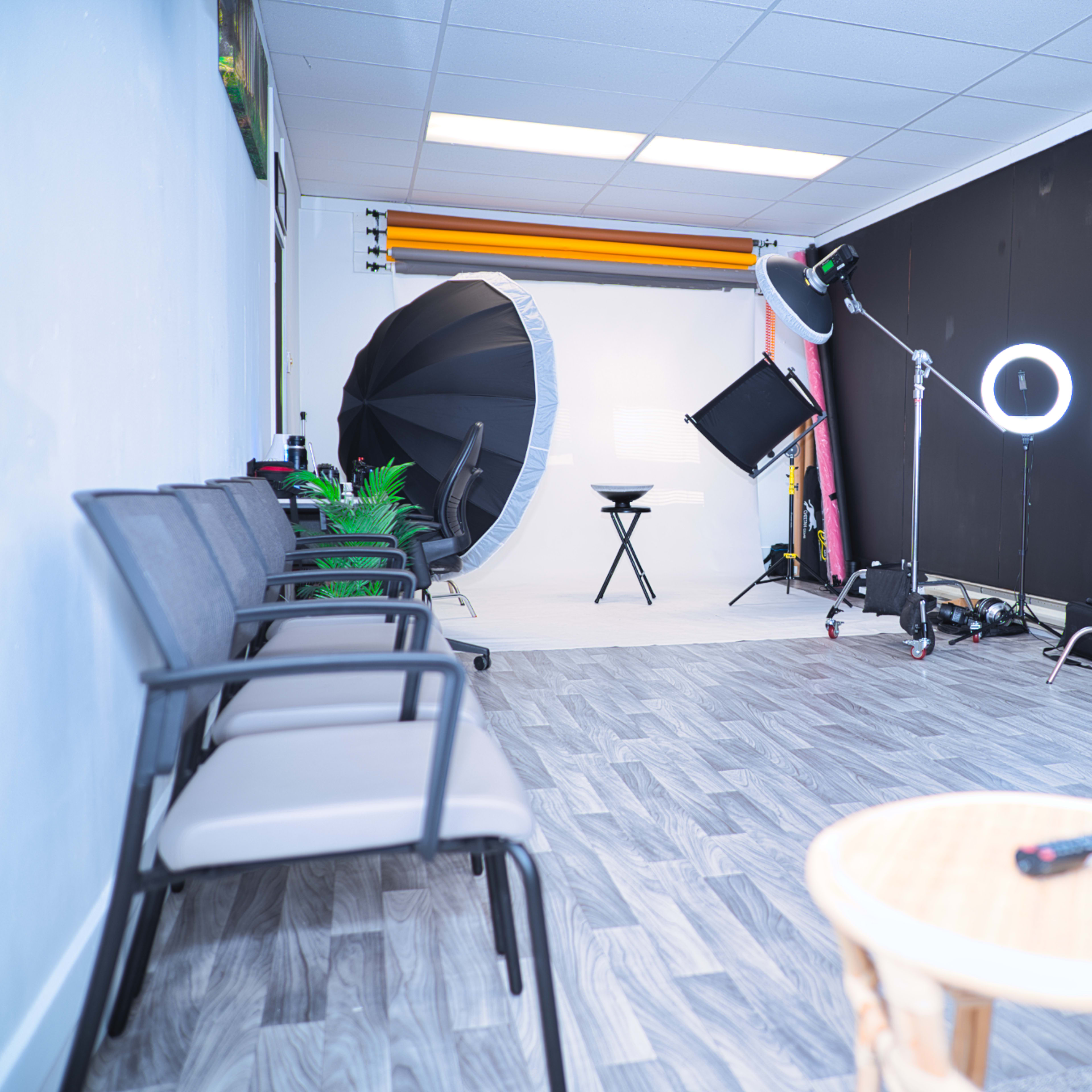 A photography studio setup with chairs on one side, a large umbrella light, a ring light, and various equipment against a black backdrop.