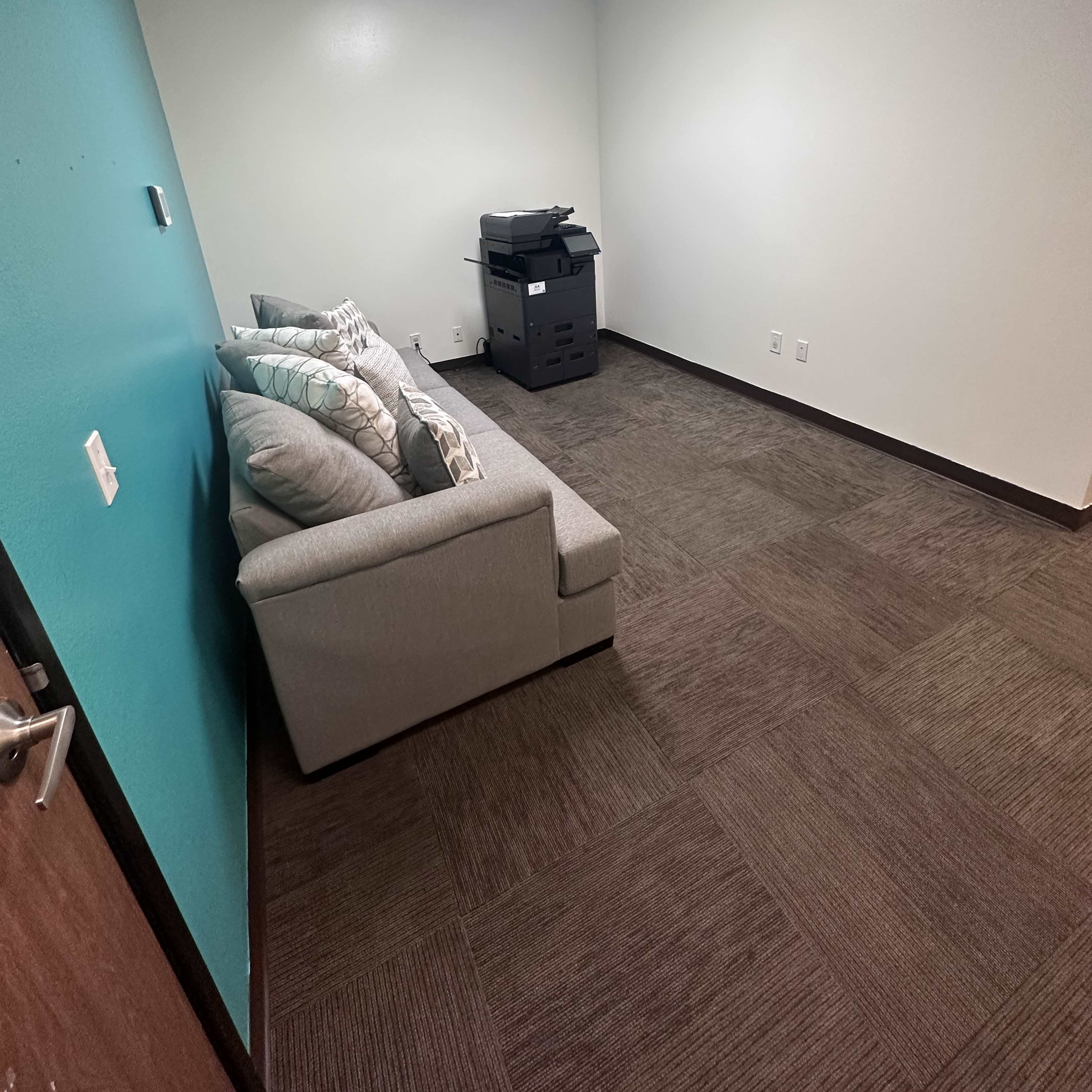 A small, empty room with a light blue wall, a gray couch with pillows, and a printer stationed in the corner.
