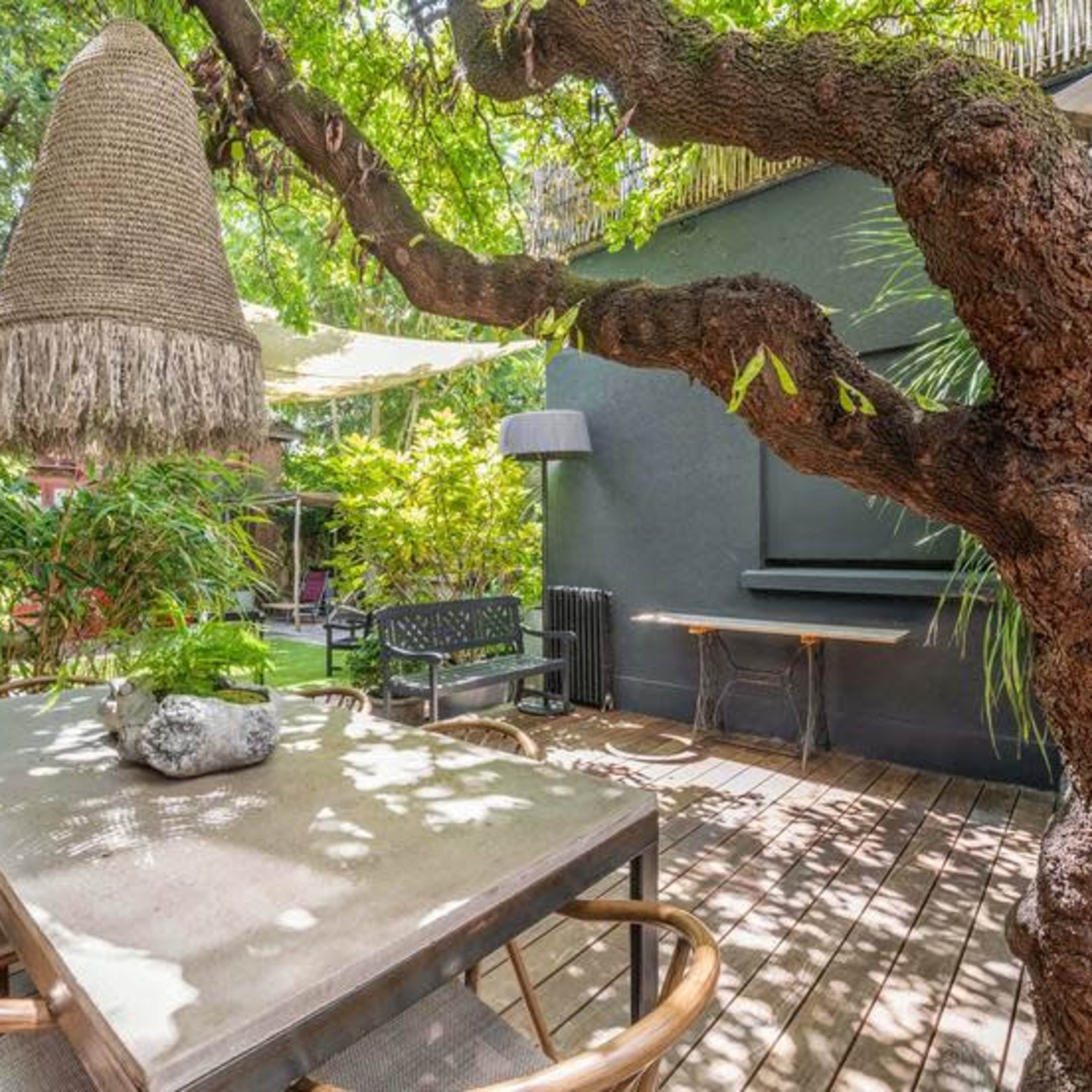 A wooden table with wicker chairs sits beneath a large tree featuring a hanging decorative light, surrounded by lush greenery.