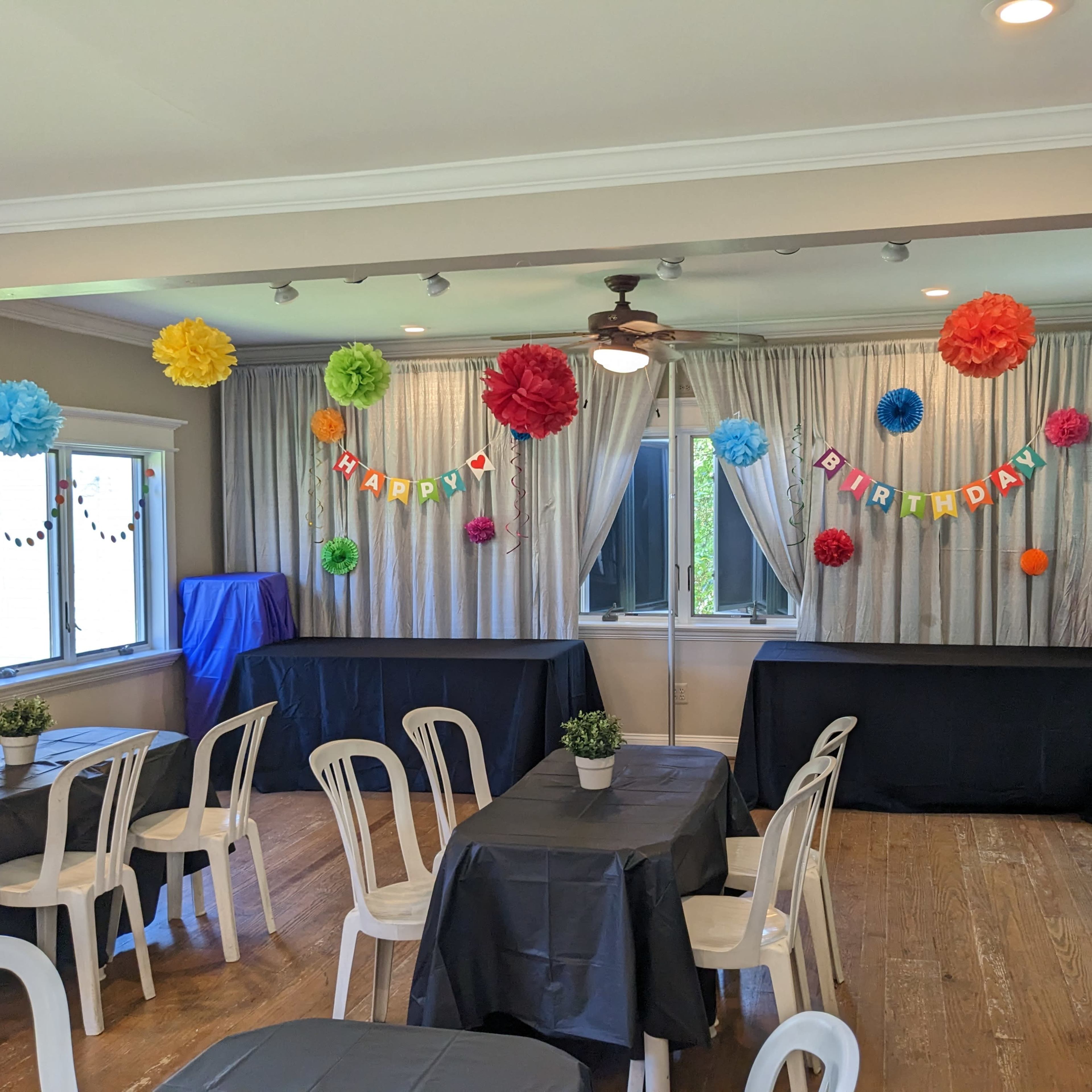 A decorated event space features colorful tissue paper flowers and a "Happy Birthday" banner, with tables and chairs arranged for a celebration.
