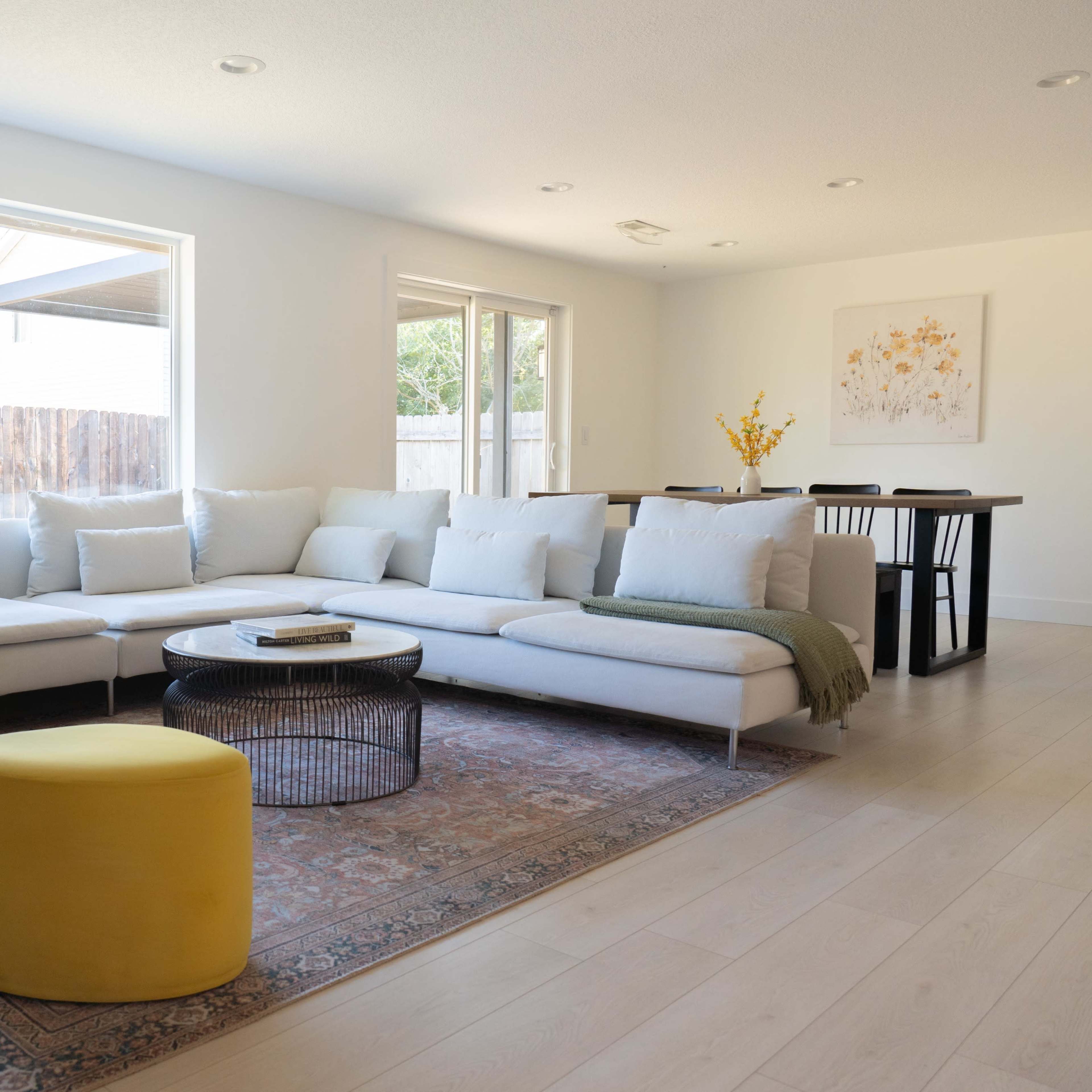 A light-filled living room features a sectional sofa with pillows, two yellow ottomans, a round coffee table, and a dining area in the background.