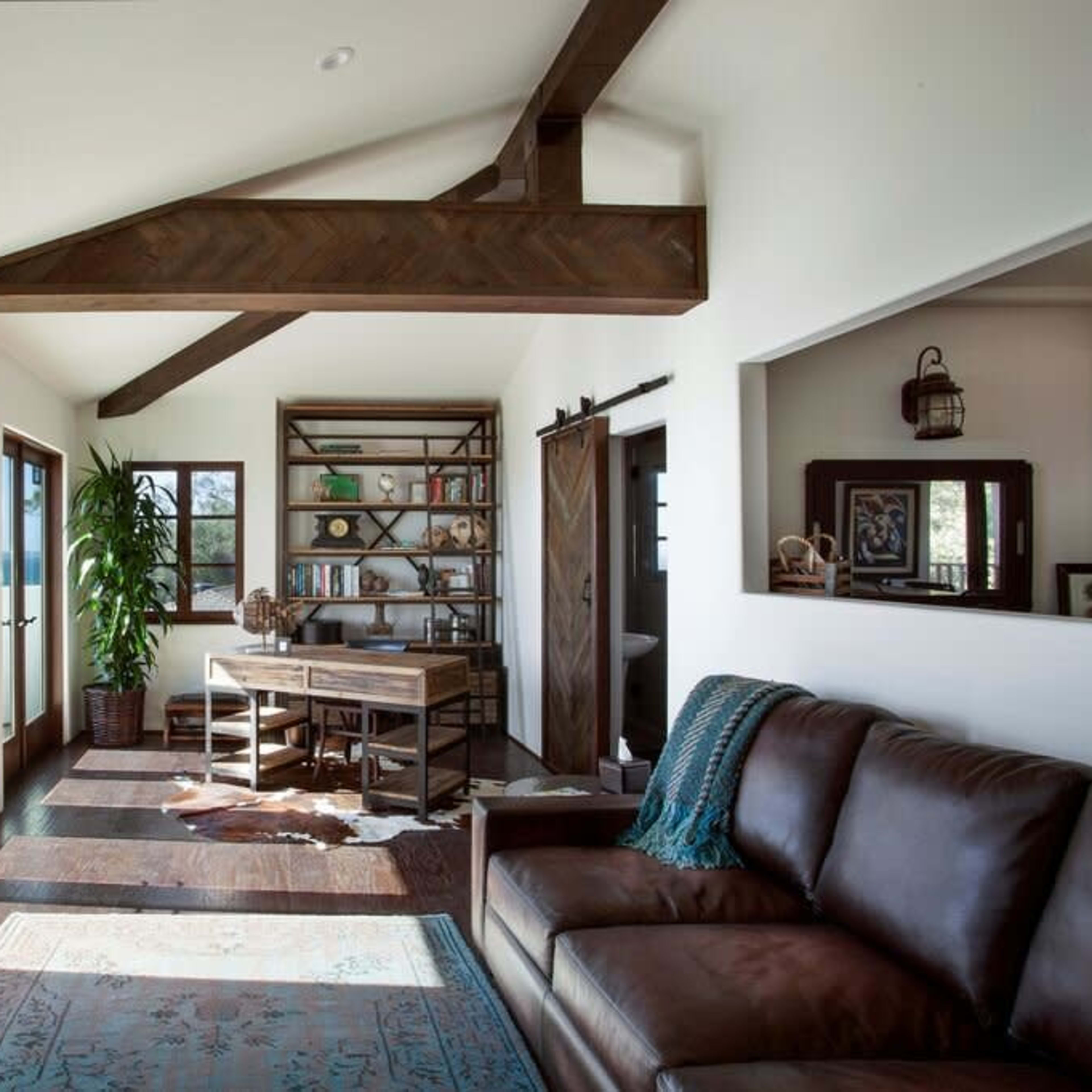A cozy living room features a brown leather sofa, a wooden desk with shelves, and large windows offering a view of the outdoors.