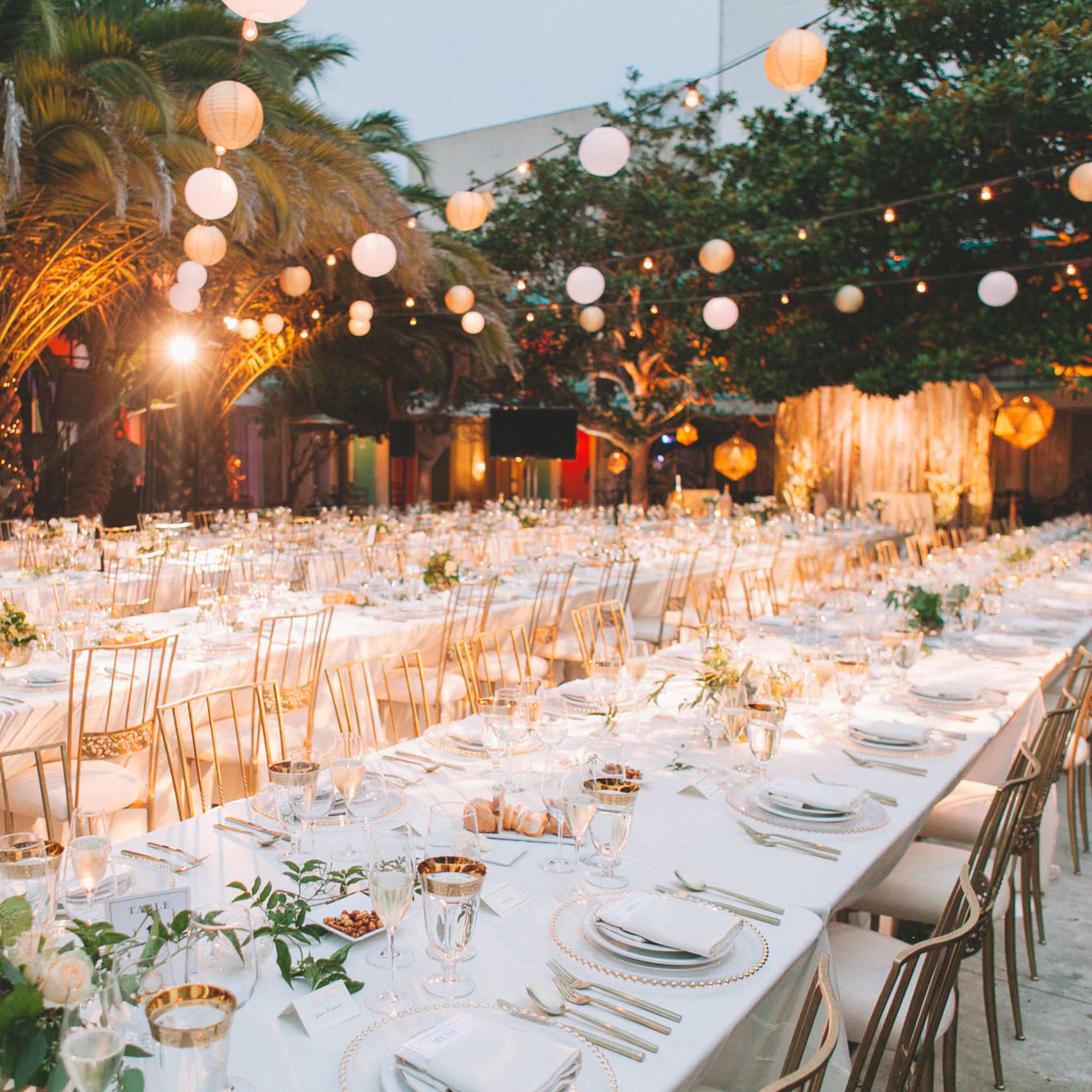 A long dining table is elegantly set under palm trees, adorned with white tablecloths, glassware, and decorative centerpieces, with string lights and lanterns illuminating the scene.