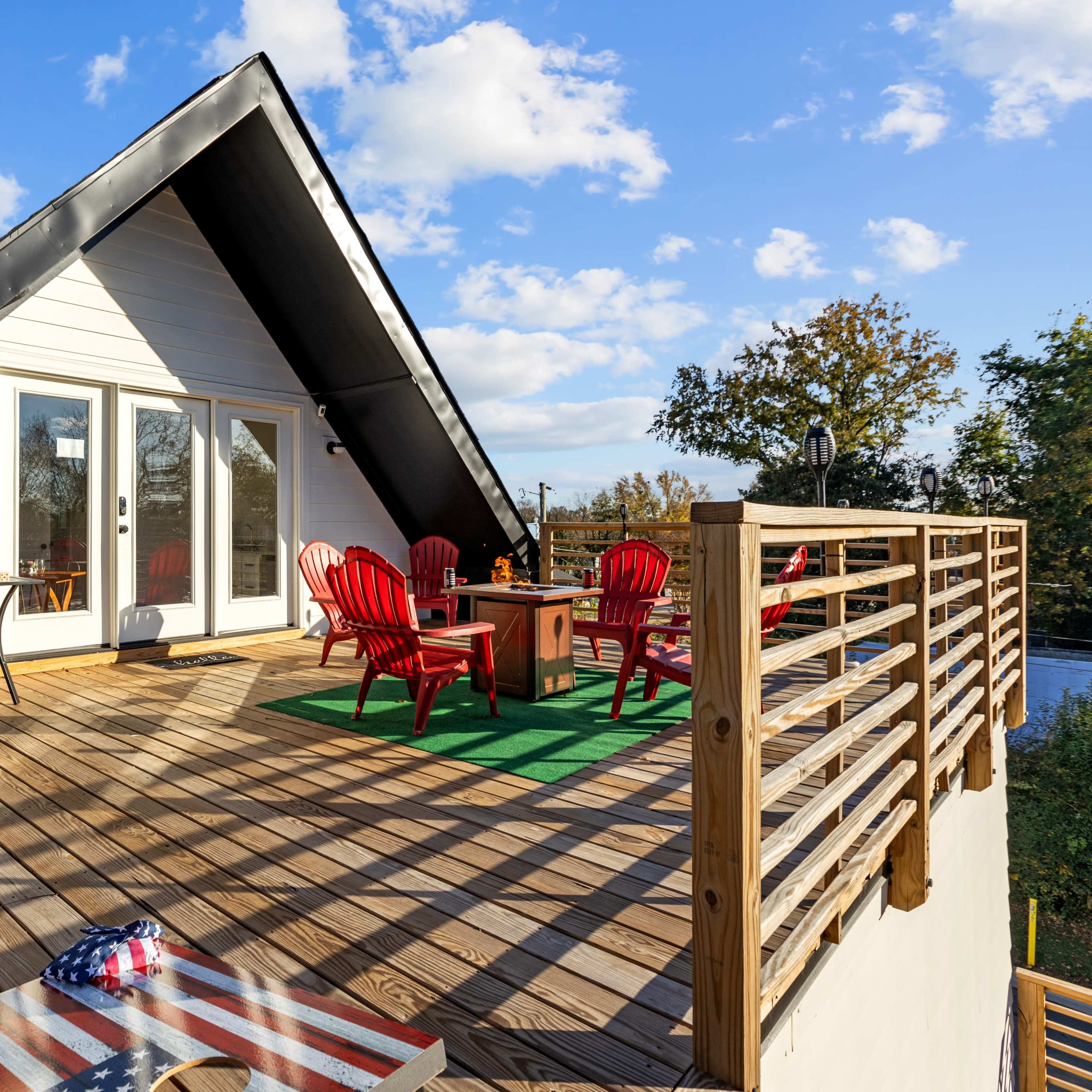 A wooden deck with red Adirondack chairs and a small table, leading to glass doors of a house beneath a sloped roof.