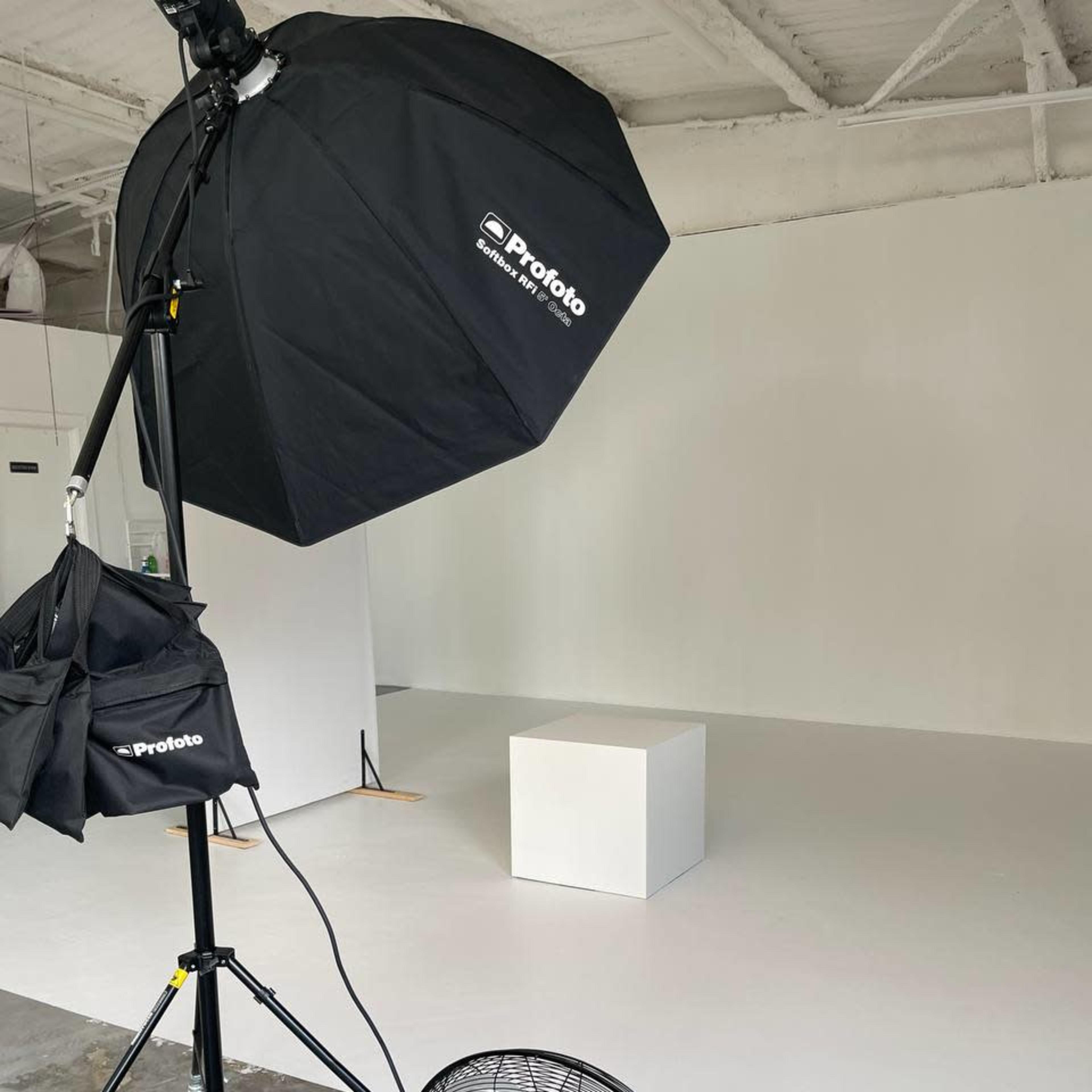 A professional photography studio features a large softbox light and a fan positioned near a white pedestal on a white backdrop.
