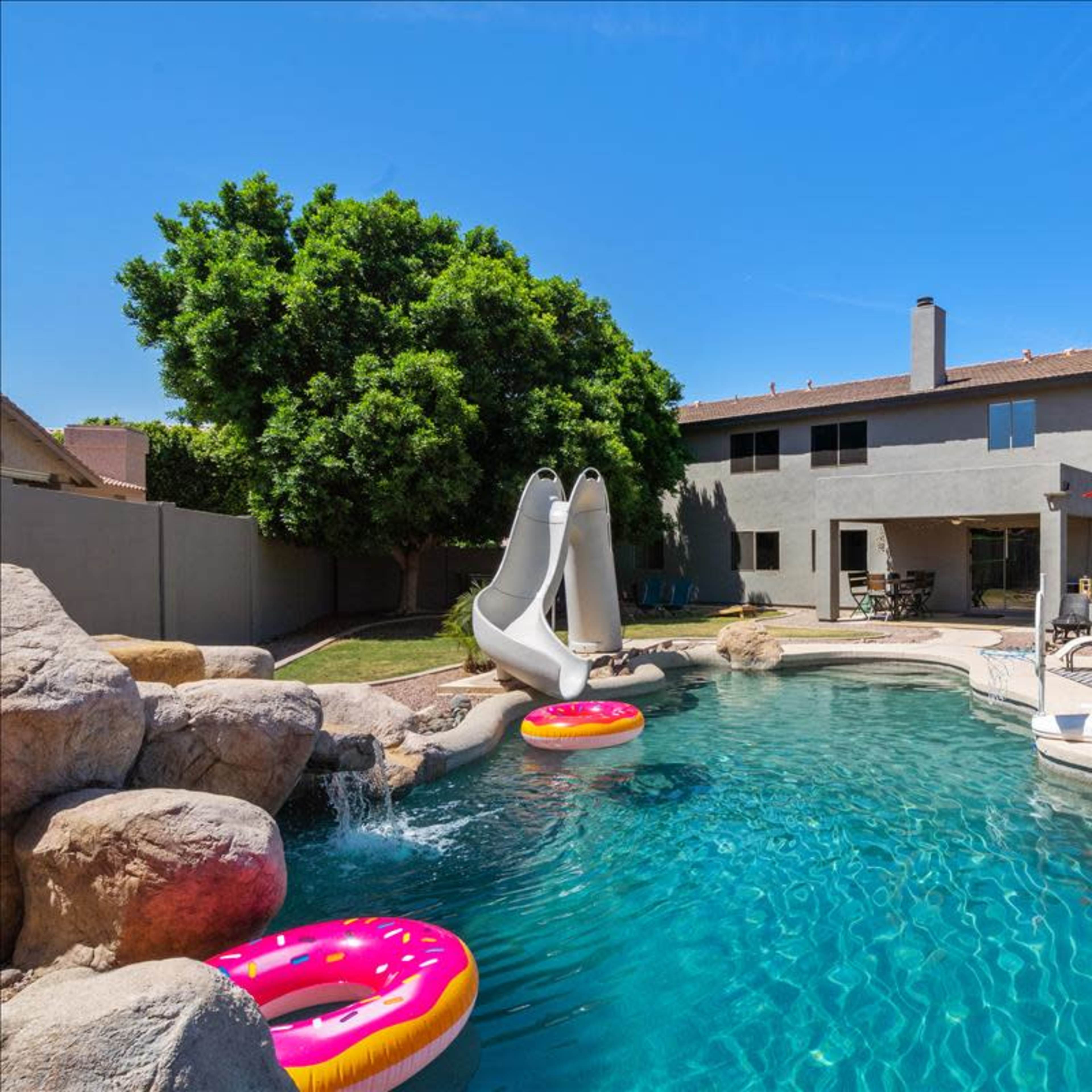 A backyard swimming pool features two water slides, inflatable rings, and a shaded seating area beside a house.