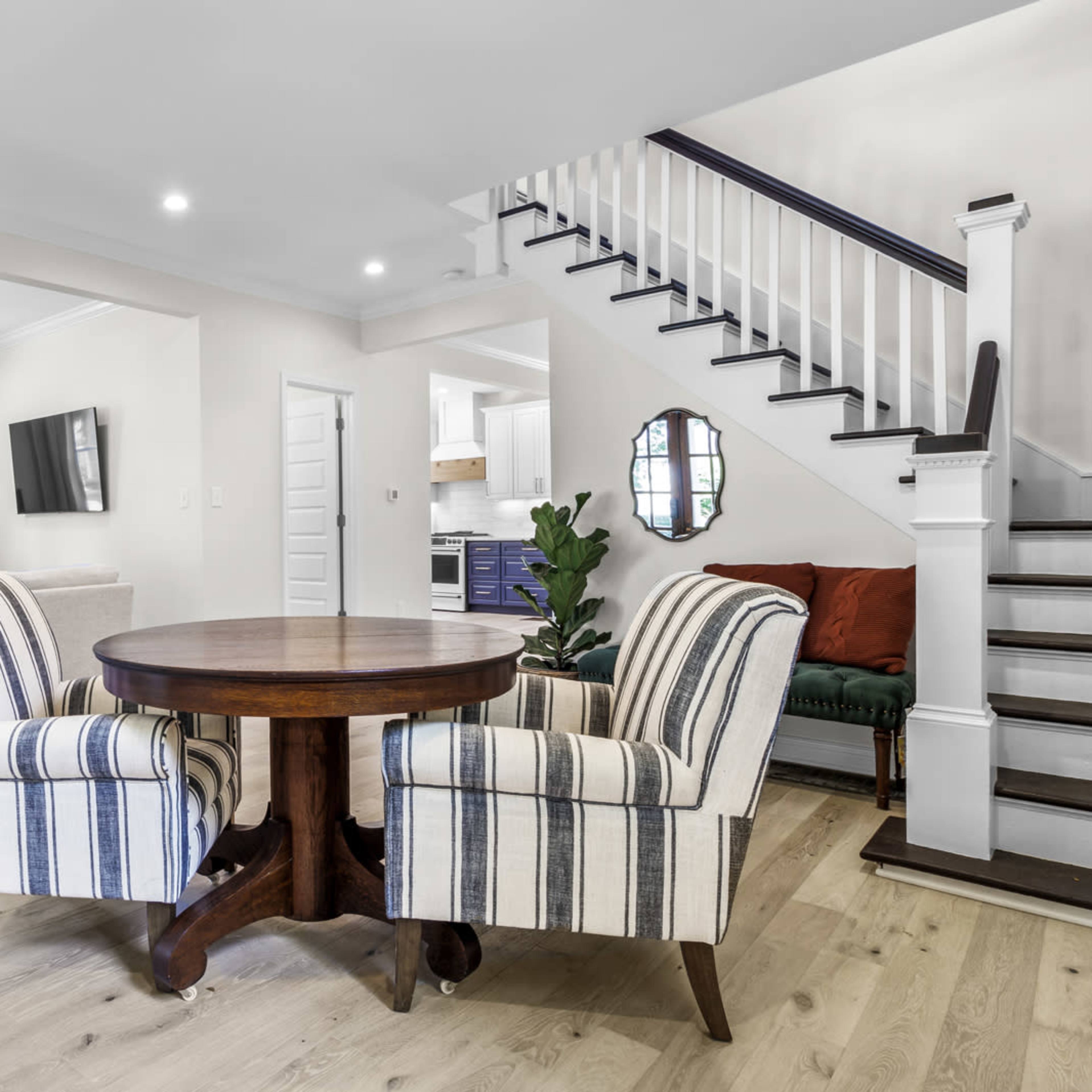 A modern interior featuring a round wooden dining table with two striped chairs, a staircase, and a living area visible in the background.