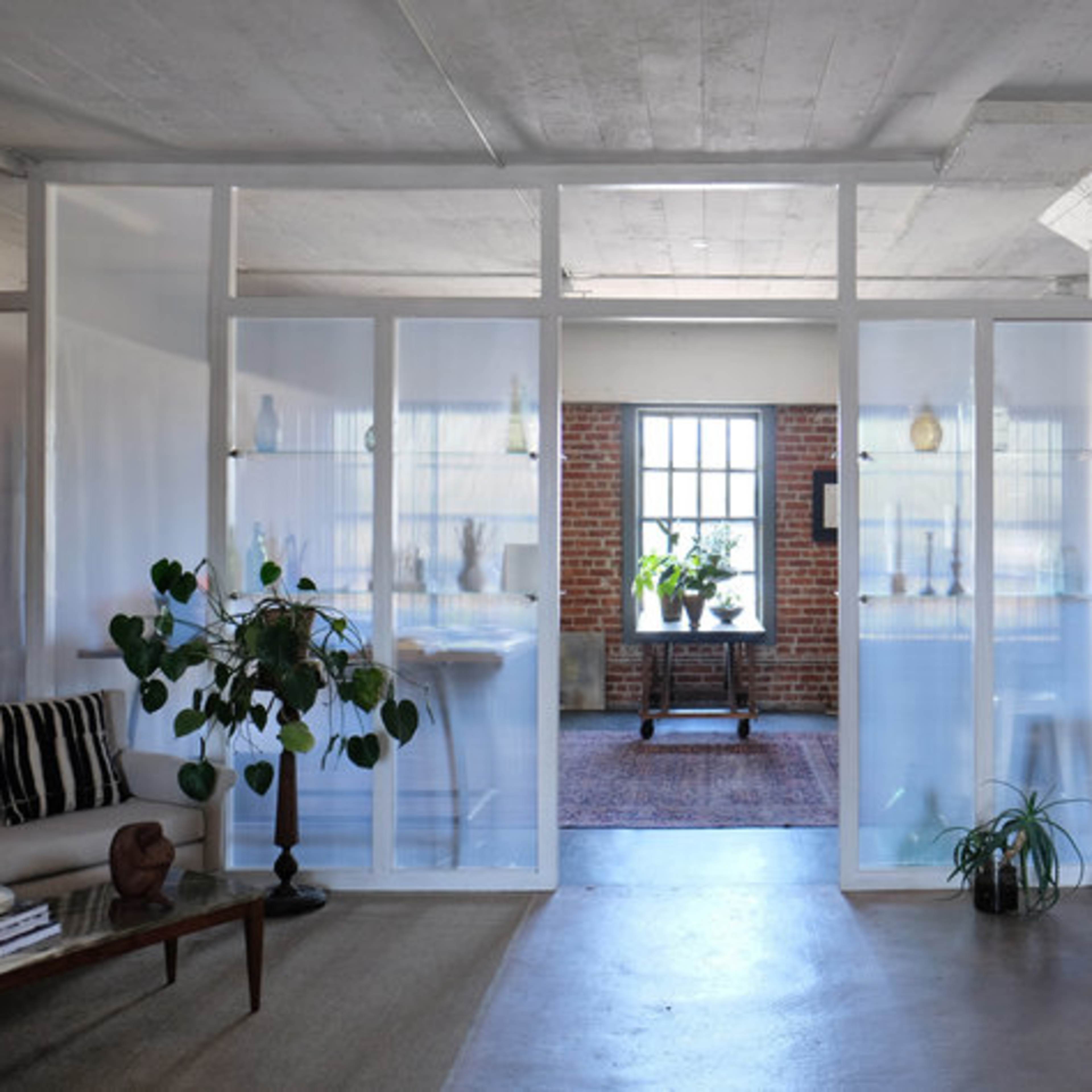 A modern, open-concept living space featuring exposed brick walls, large windows, and minimalist furniture, separated by transparent partition walls.