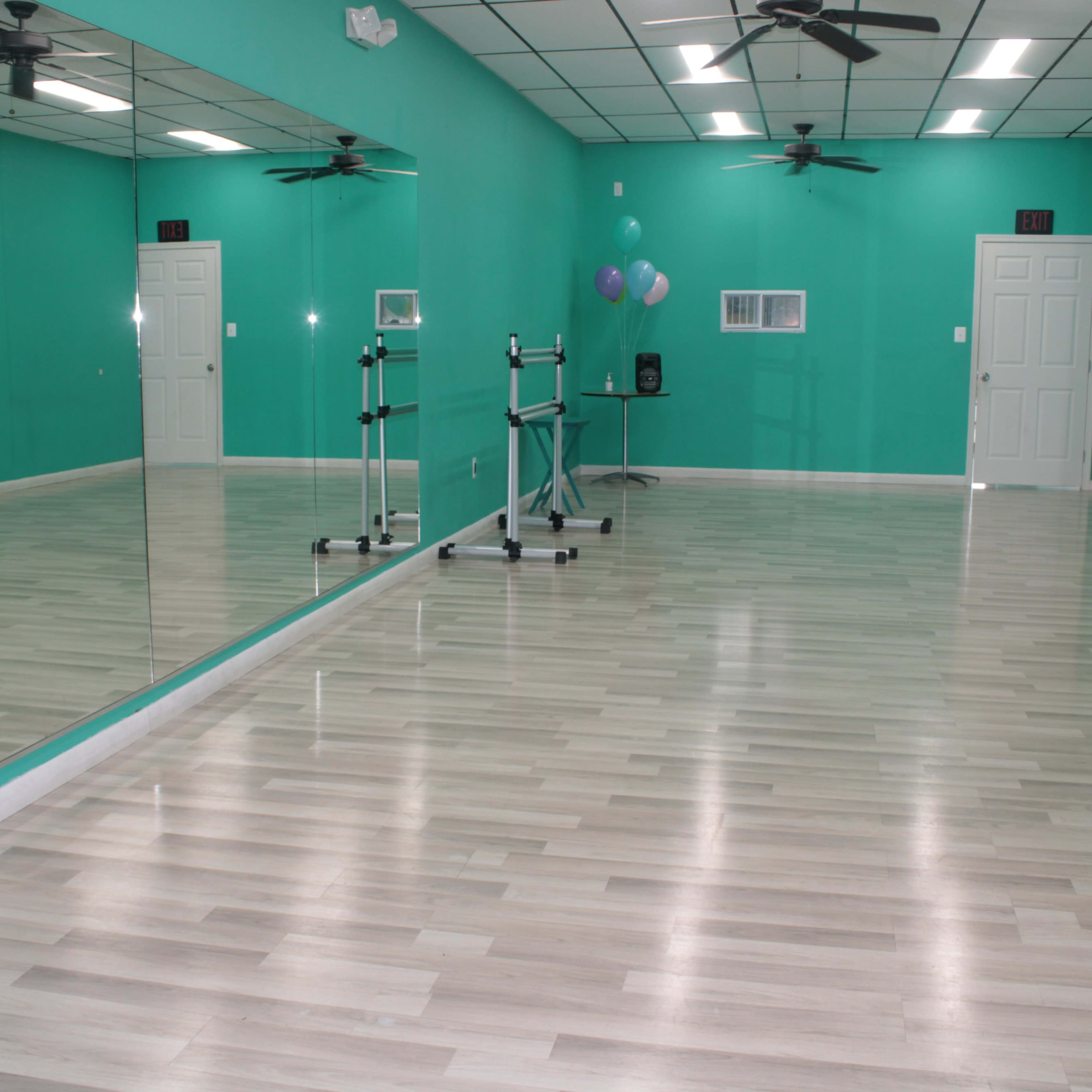 A bright, empty dance studio with teal walls, wooden floors, and large mirrors reflecting the space.