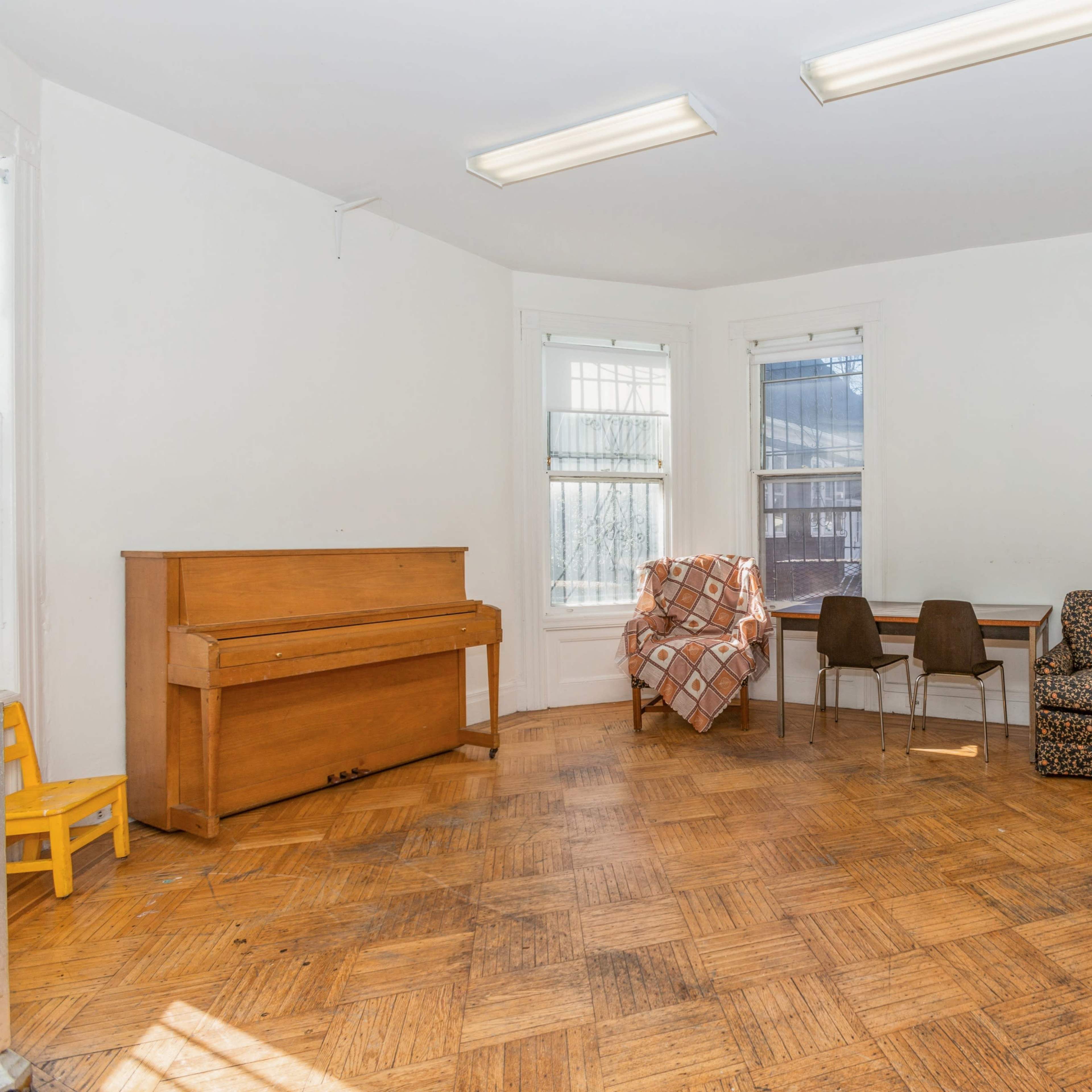 A sparse room with a piano, two chairs, and a patterned armchair surrounded by large windows and a fireplace.
