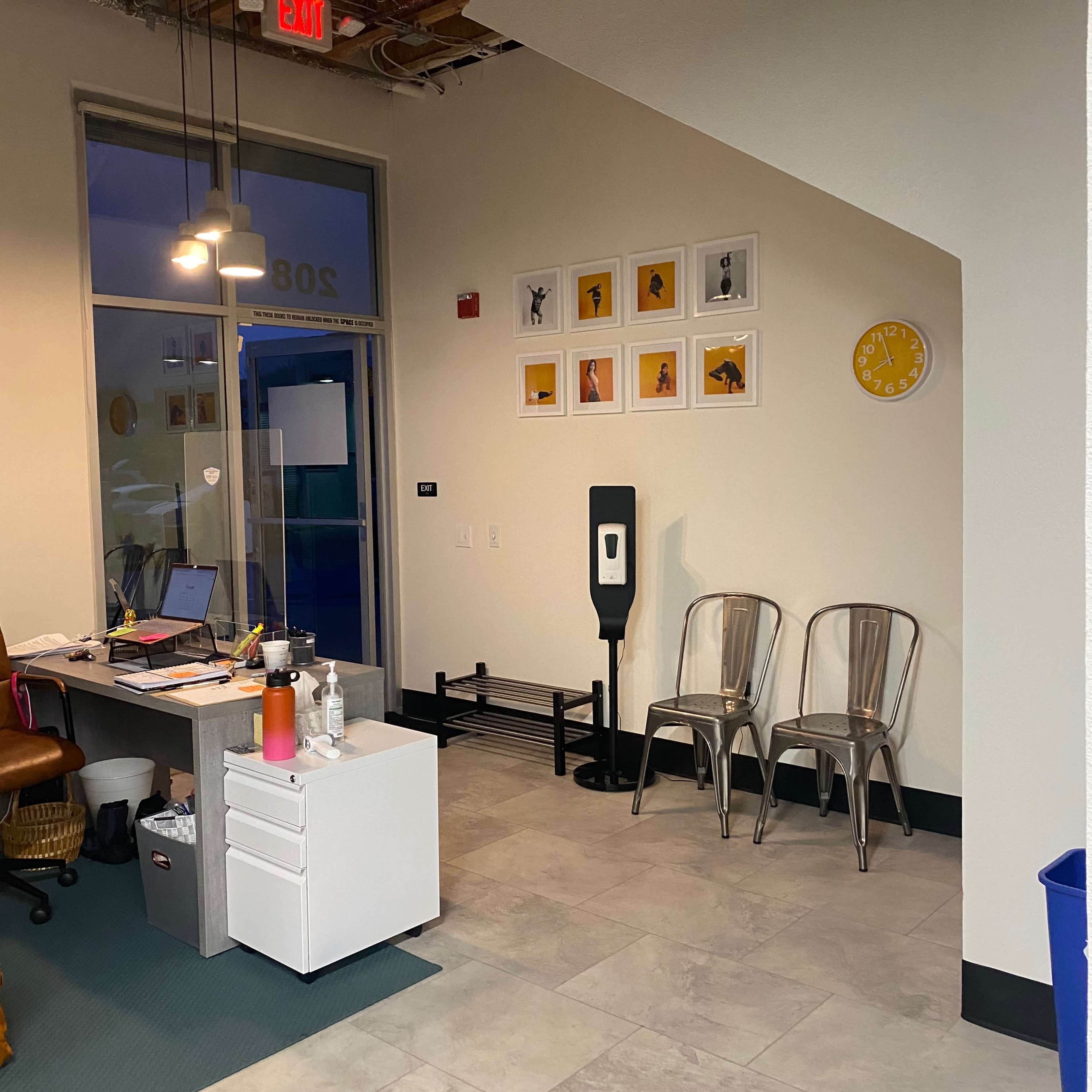 An office space featuring a desk with a computer and a filing cabinet, along with two metal chairs and a wall decorated with framed art.