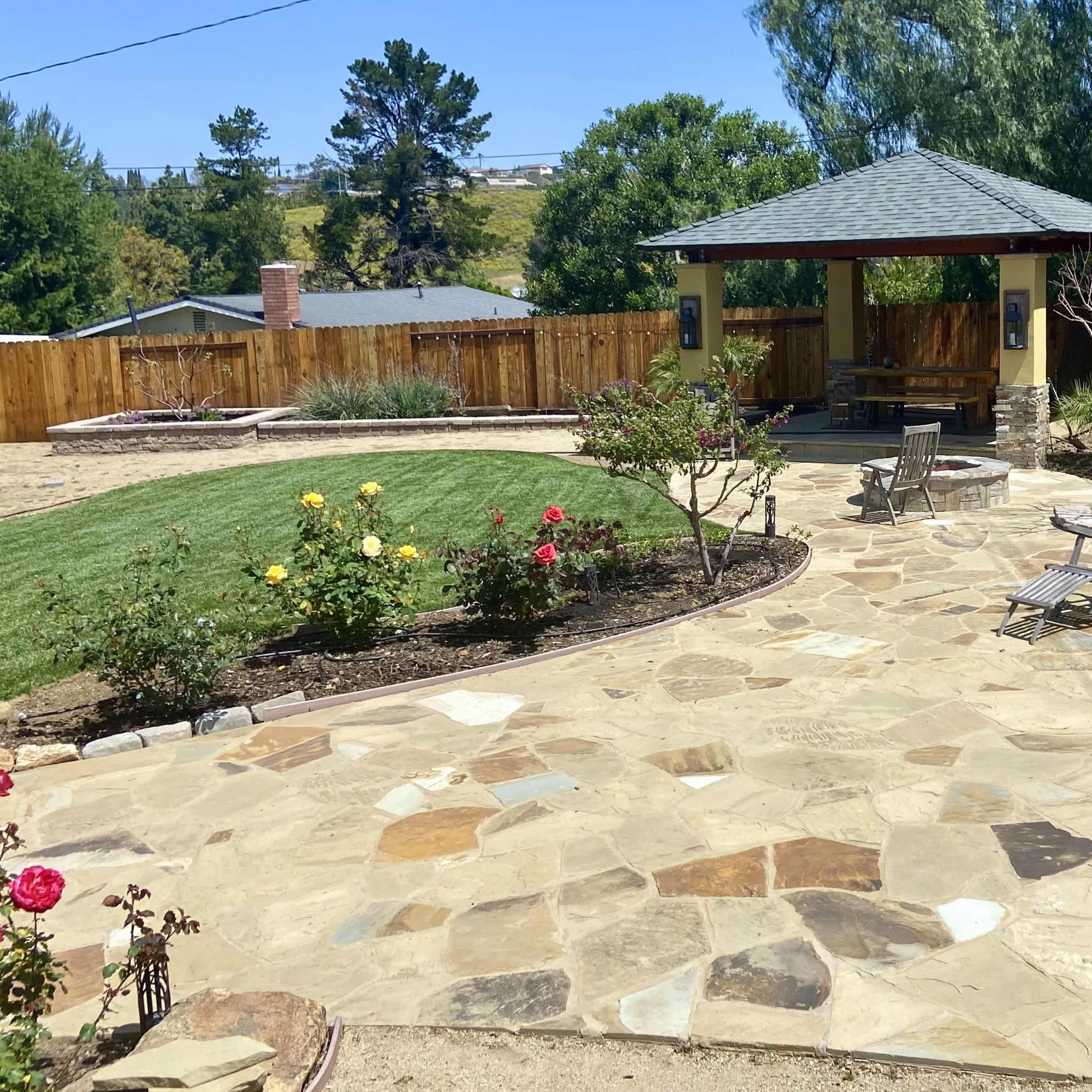 A landscaped backyard with a stone patio, a gazebo, flower beds with roses, and a grassy area surrounded by a wooden fence.