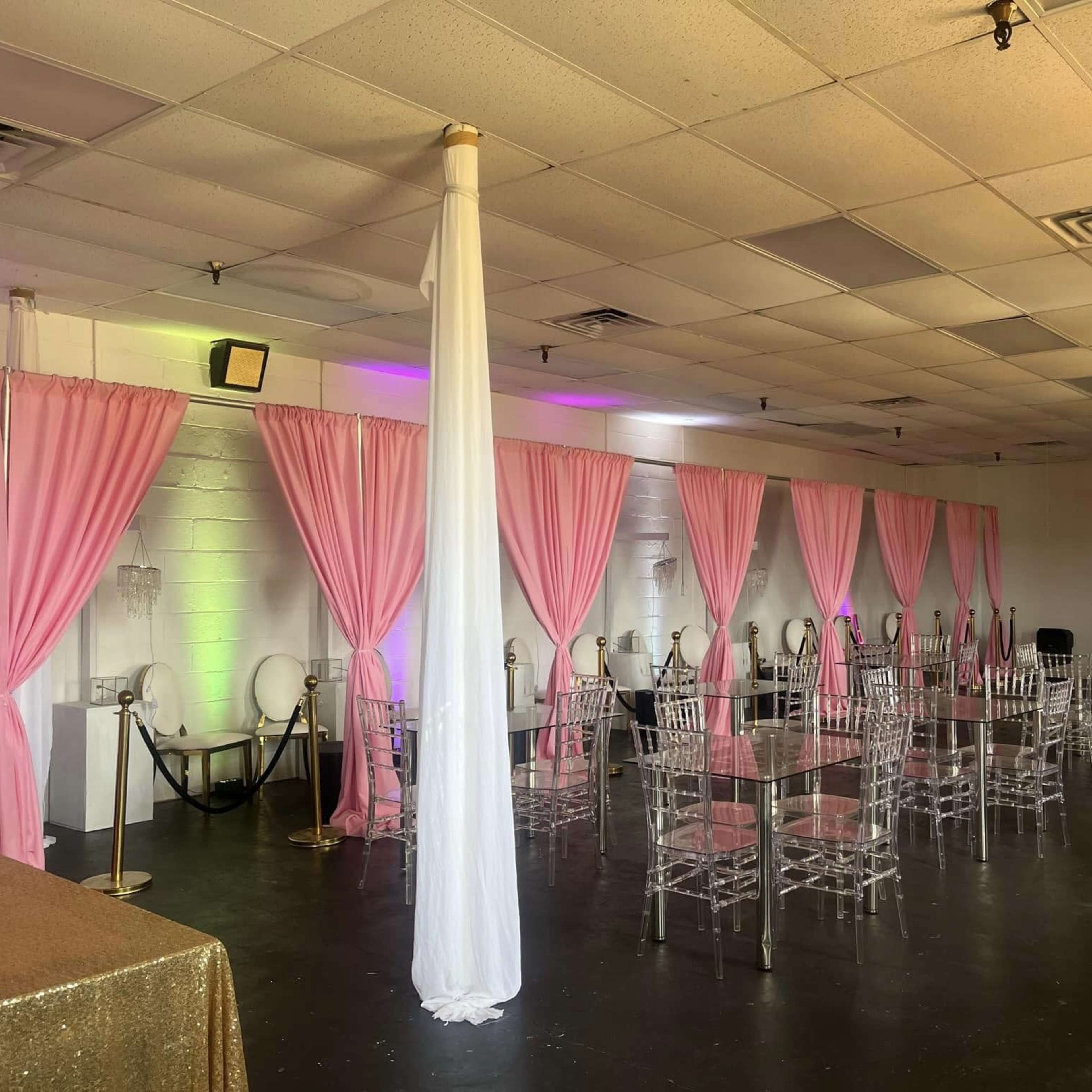 An event space with pink drapes, clear acrylic chairs, and a gold tablecloth, arranged for a gathering.