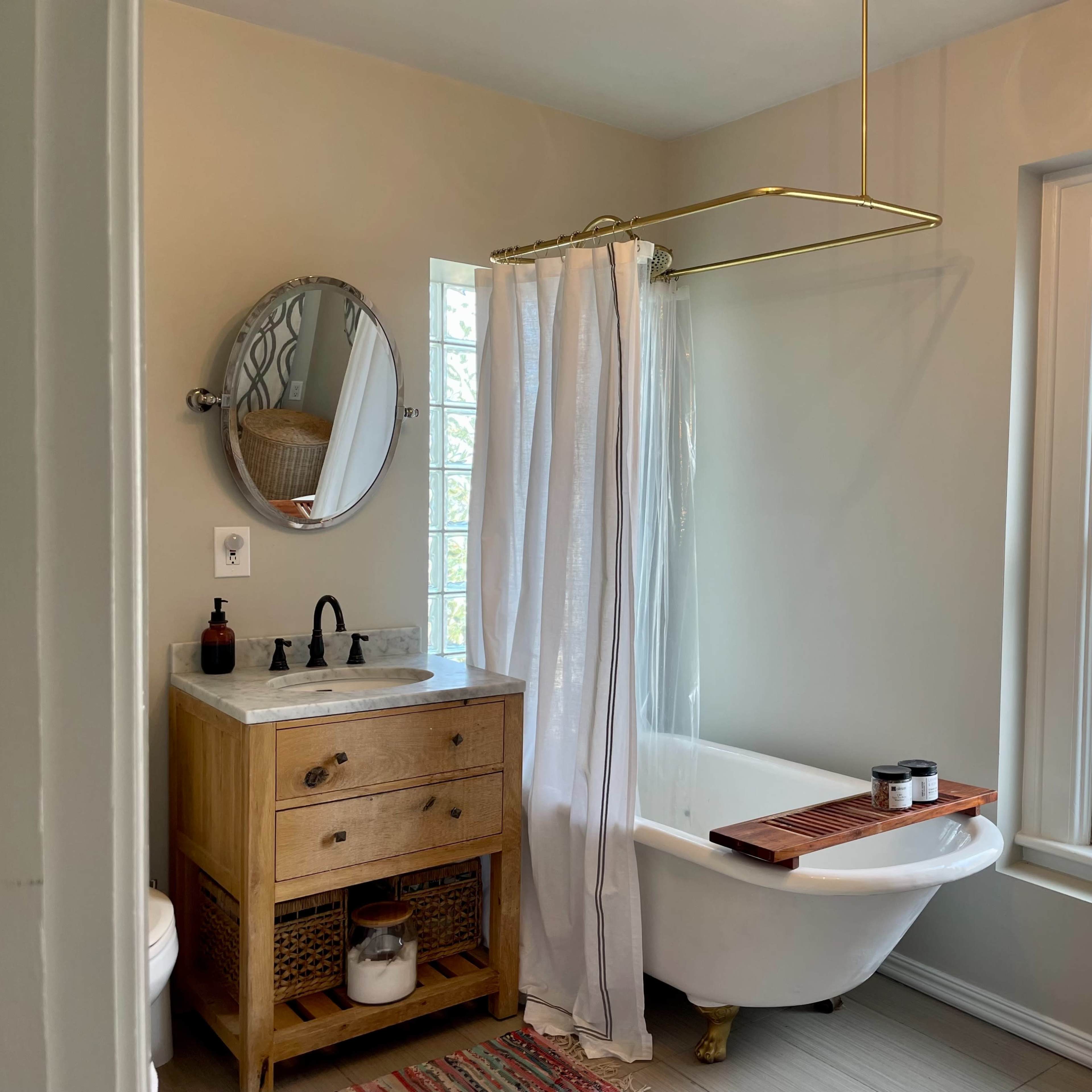 A modern bathroom featuring a clawfoot tub with a wooden bath tray, a circular mirror above a wooden vanity, and a window allowing natural light.
