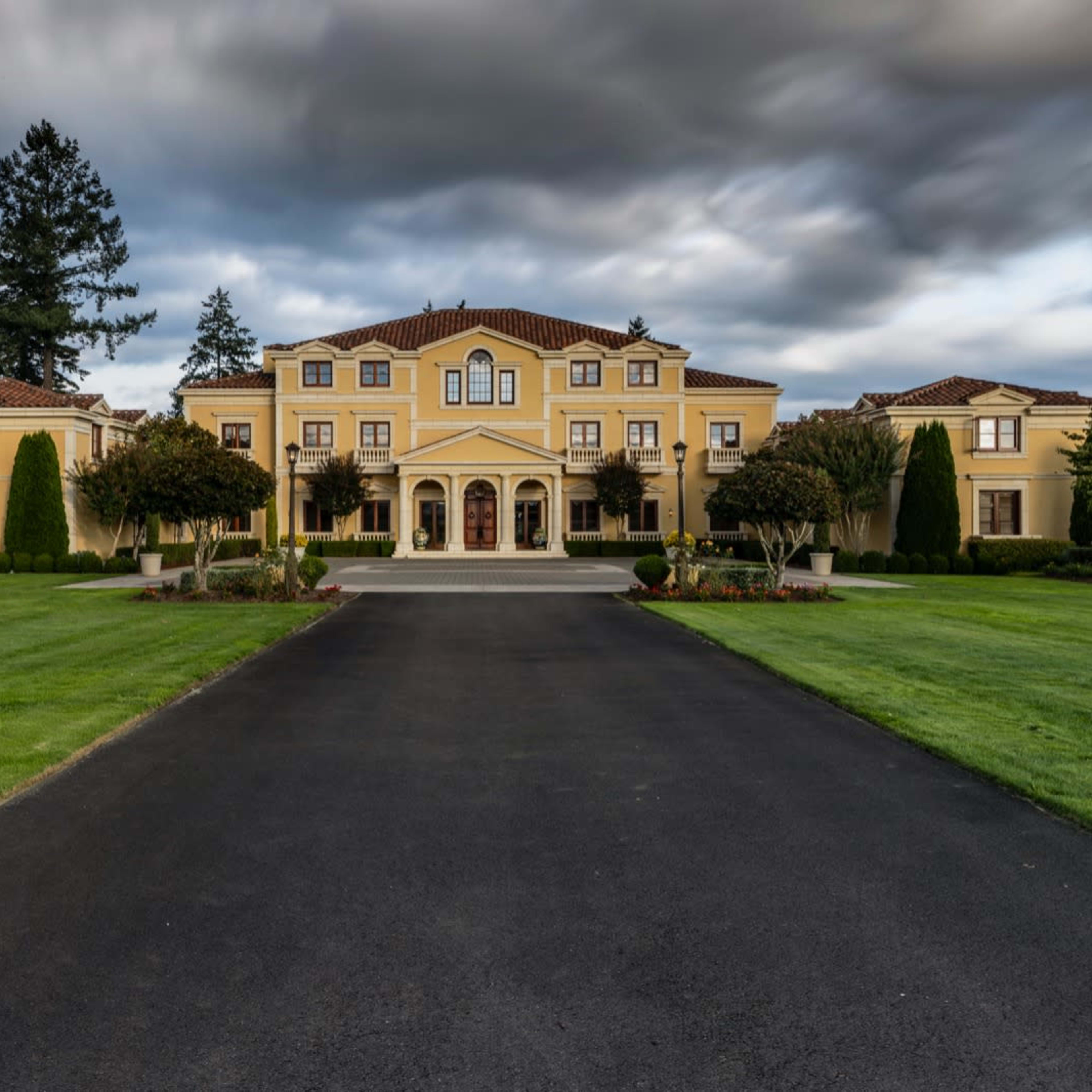 A large, yellow mansion with a tiled roof is set on manicured lawns and accessed by a long, paved driveway.