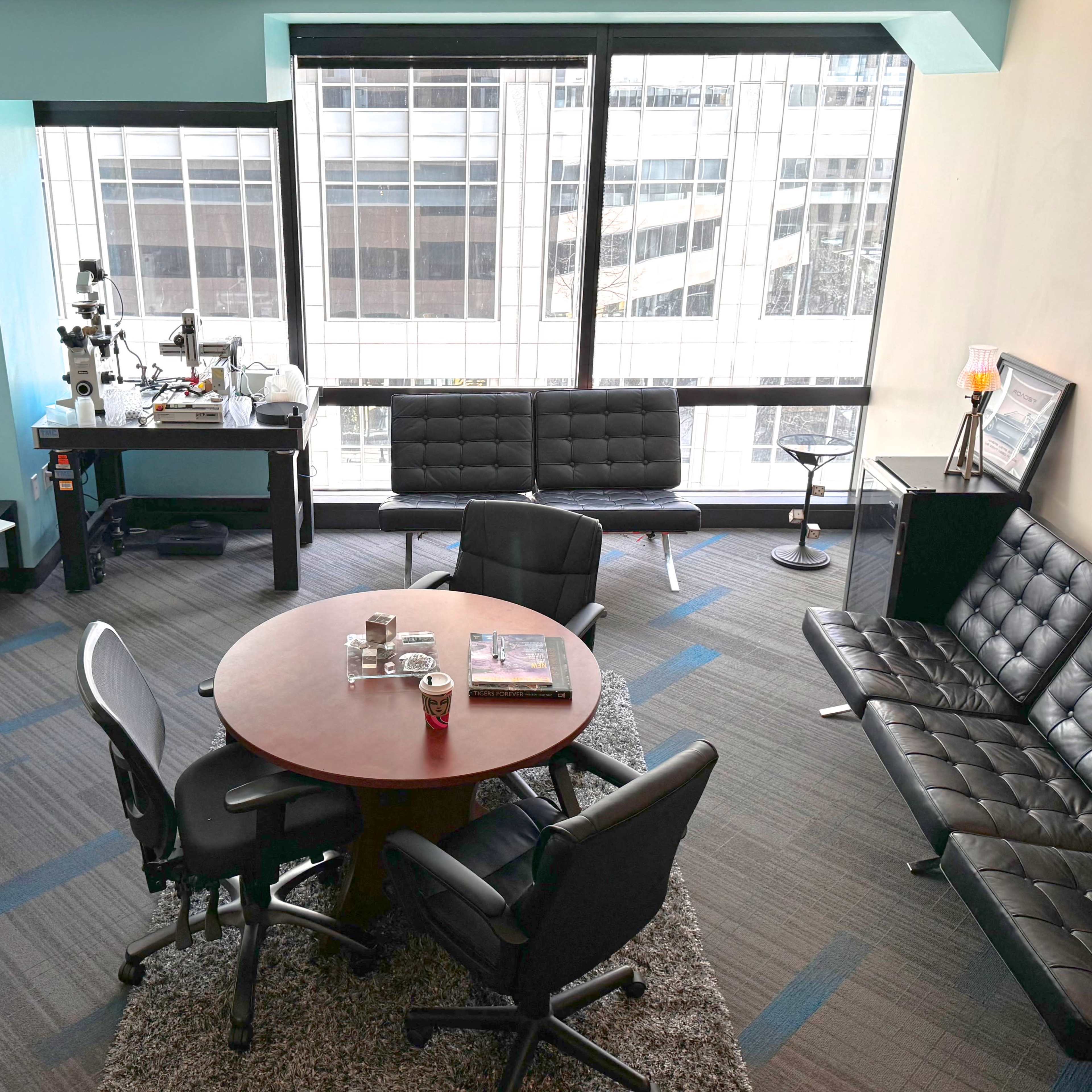 A modern office space with a round table surrounded by chairs, a black couch, a desk with equipment, and large windows overlooking a cityscape.
