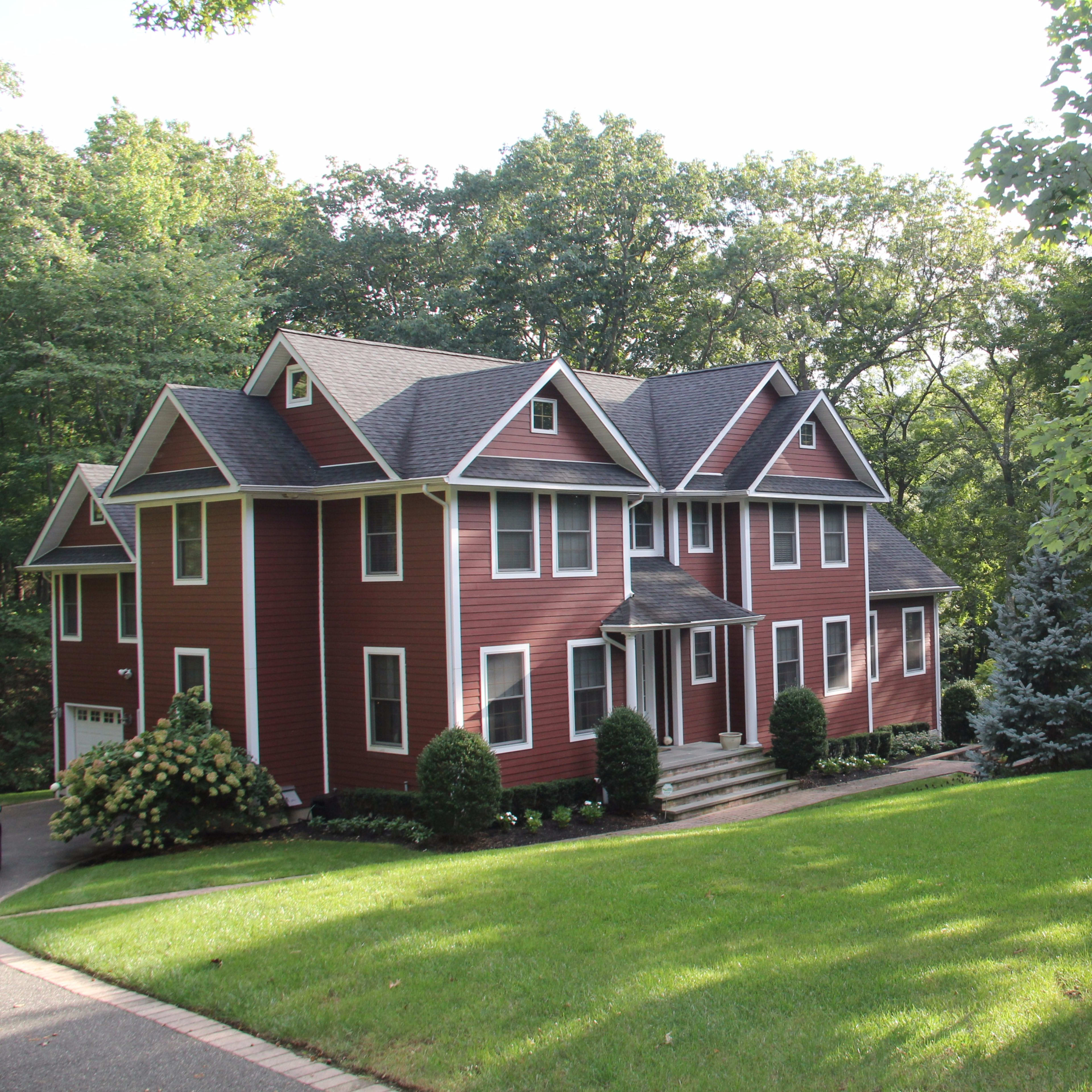 A large red house with multiple gabled roofs sits on a well-manicured lawn surrounded by trees.