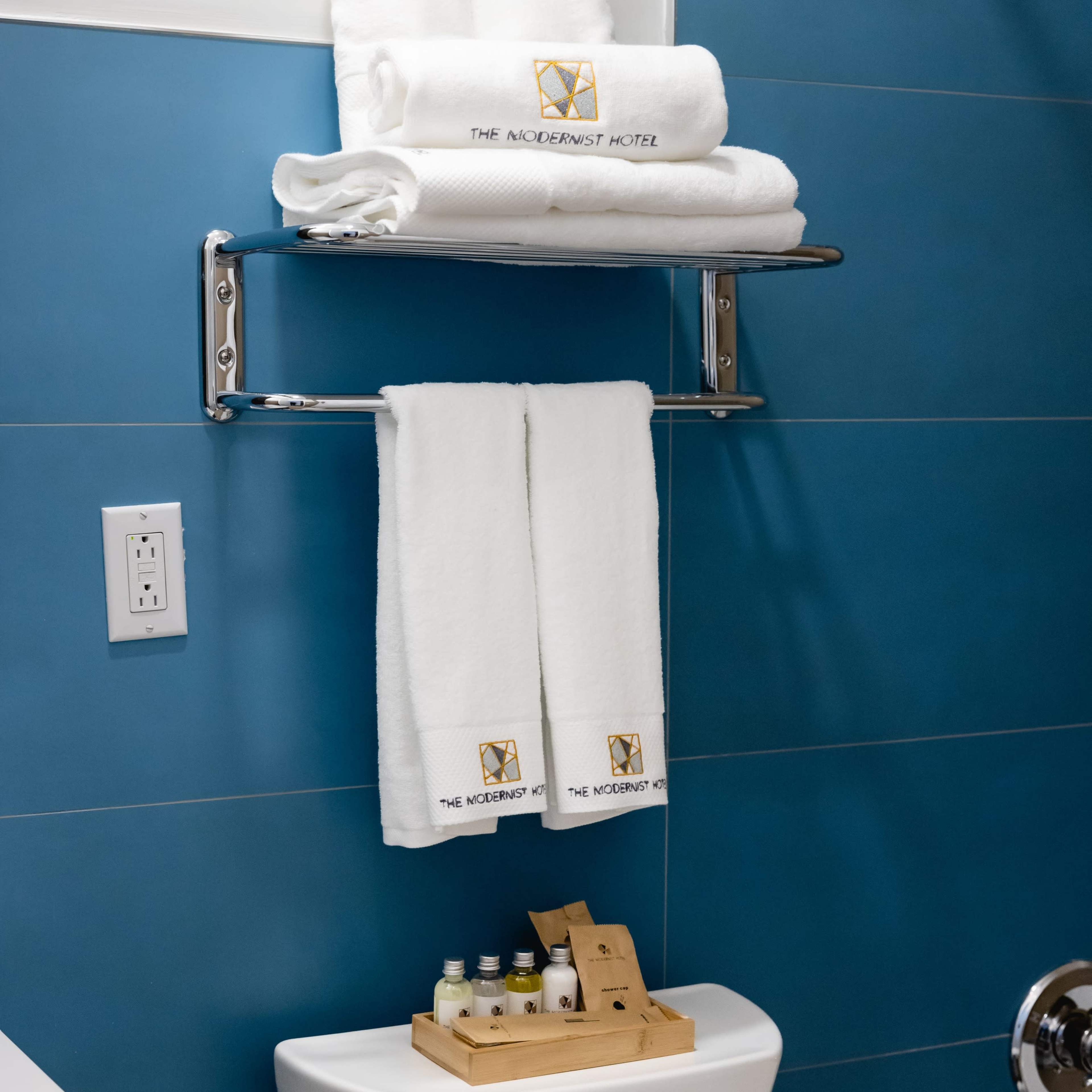 A neatly arranged bathroom displays white towels on a chrome rack above a toilet, accompanied by a wooden tray of miniature toiletries.