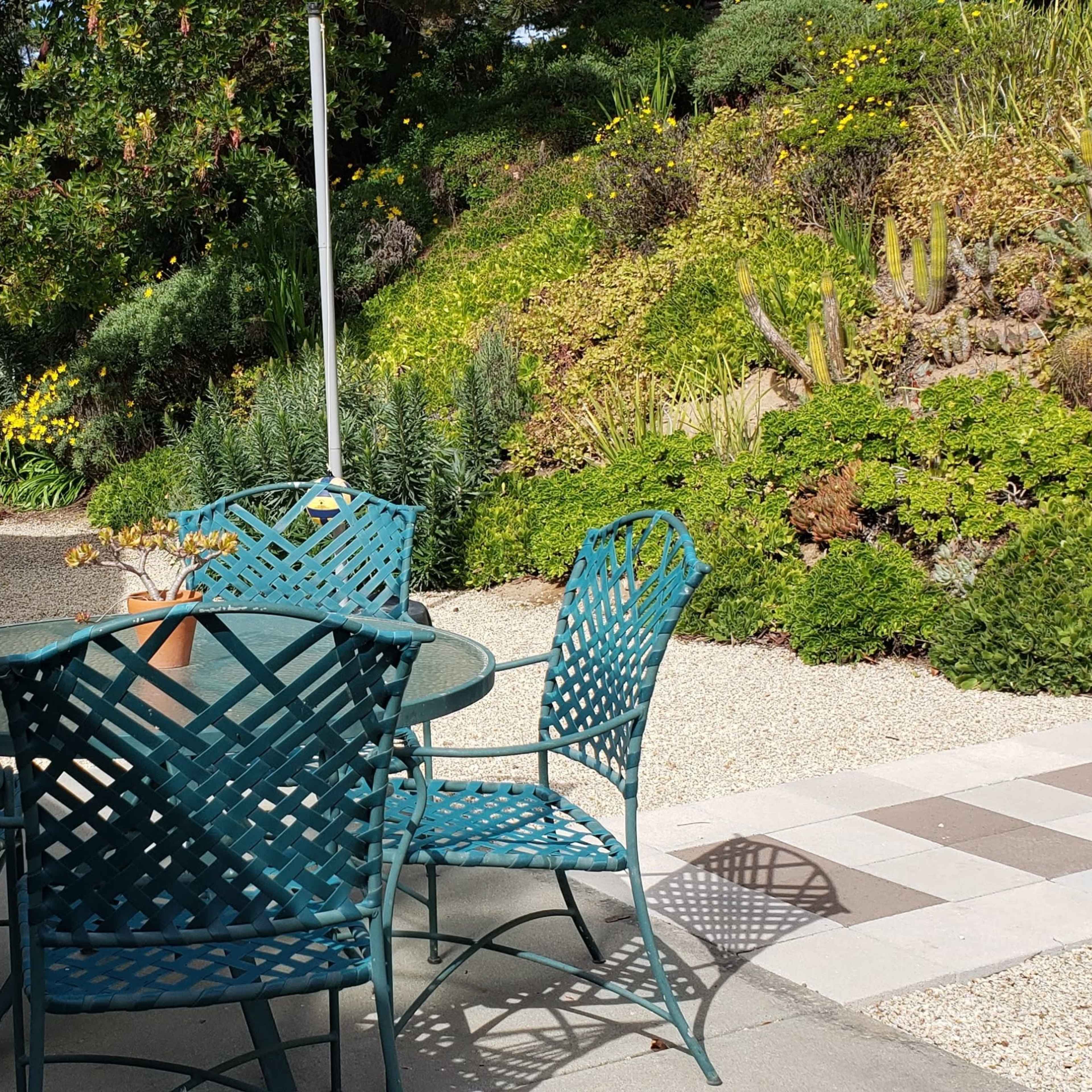 A patio area features a round glass table surrounded by four teal metal chairs, situated beside a landscaped garden with varied plants and a pathway.