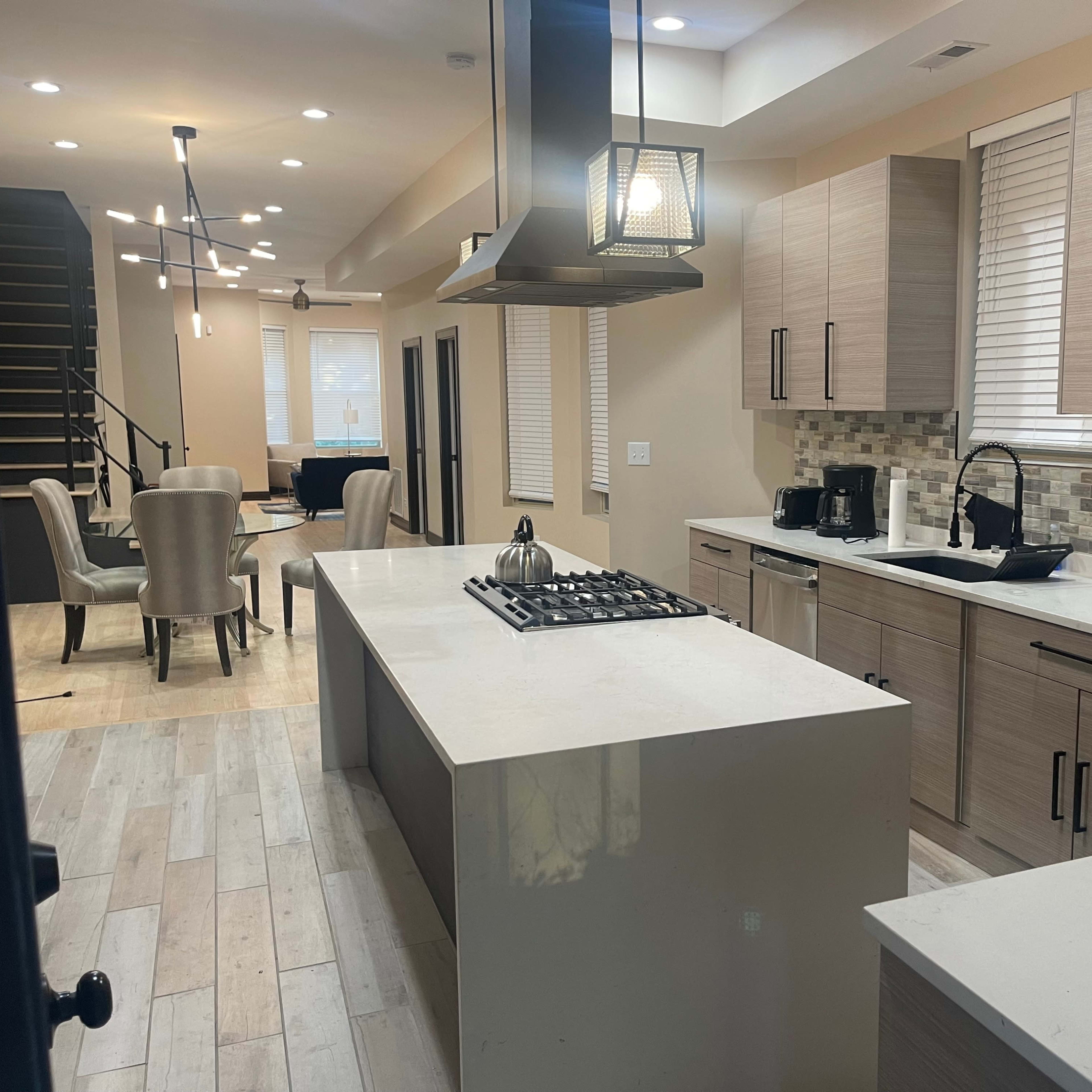 A modern kitchen with light wood cabinetry, a large island with a stovetop, and an open layout leading into a dining area with upholstered chairs.