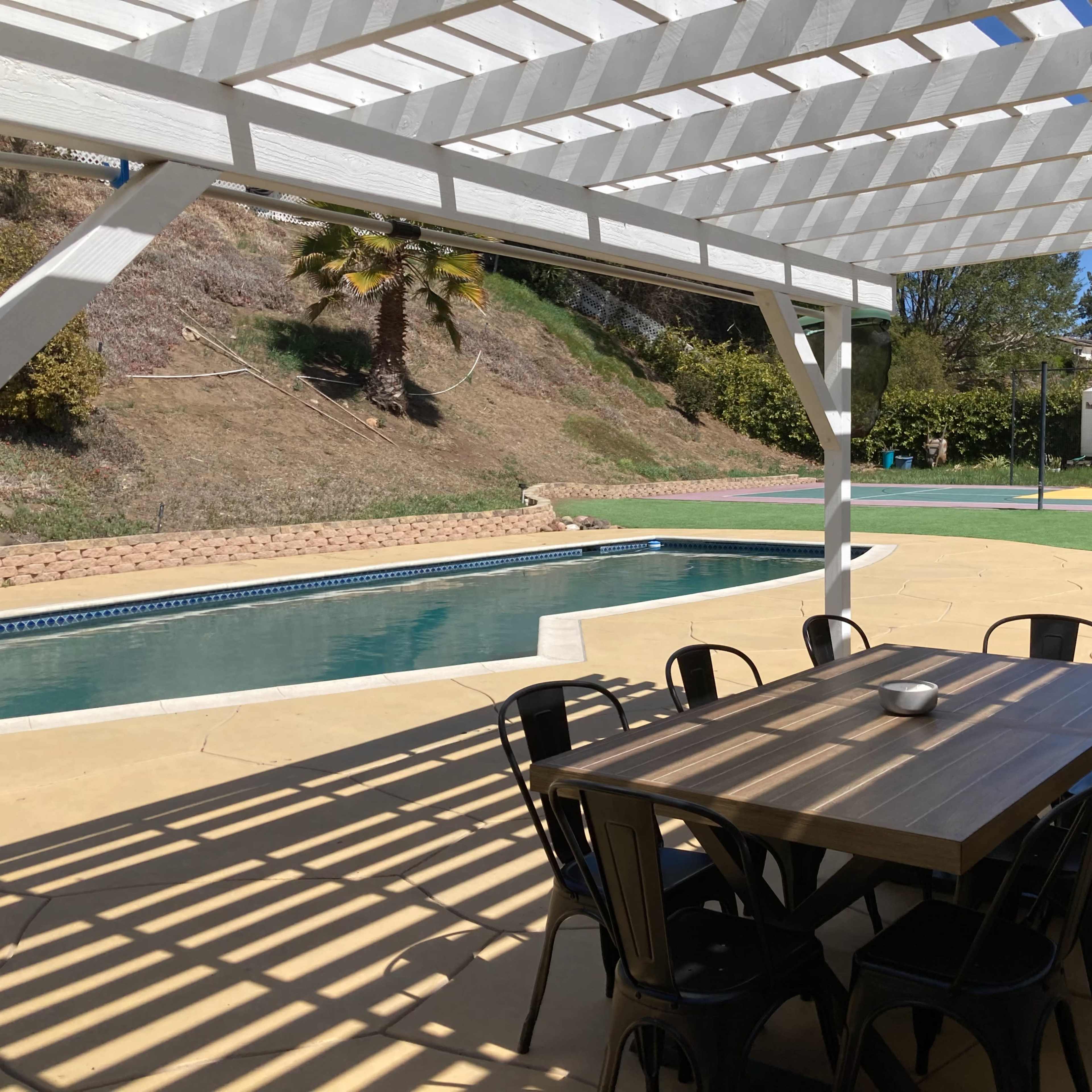 A backyard patio with a dining table and chairs beside a swimming pool and a basketball court in the background.