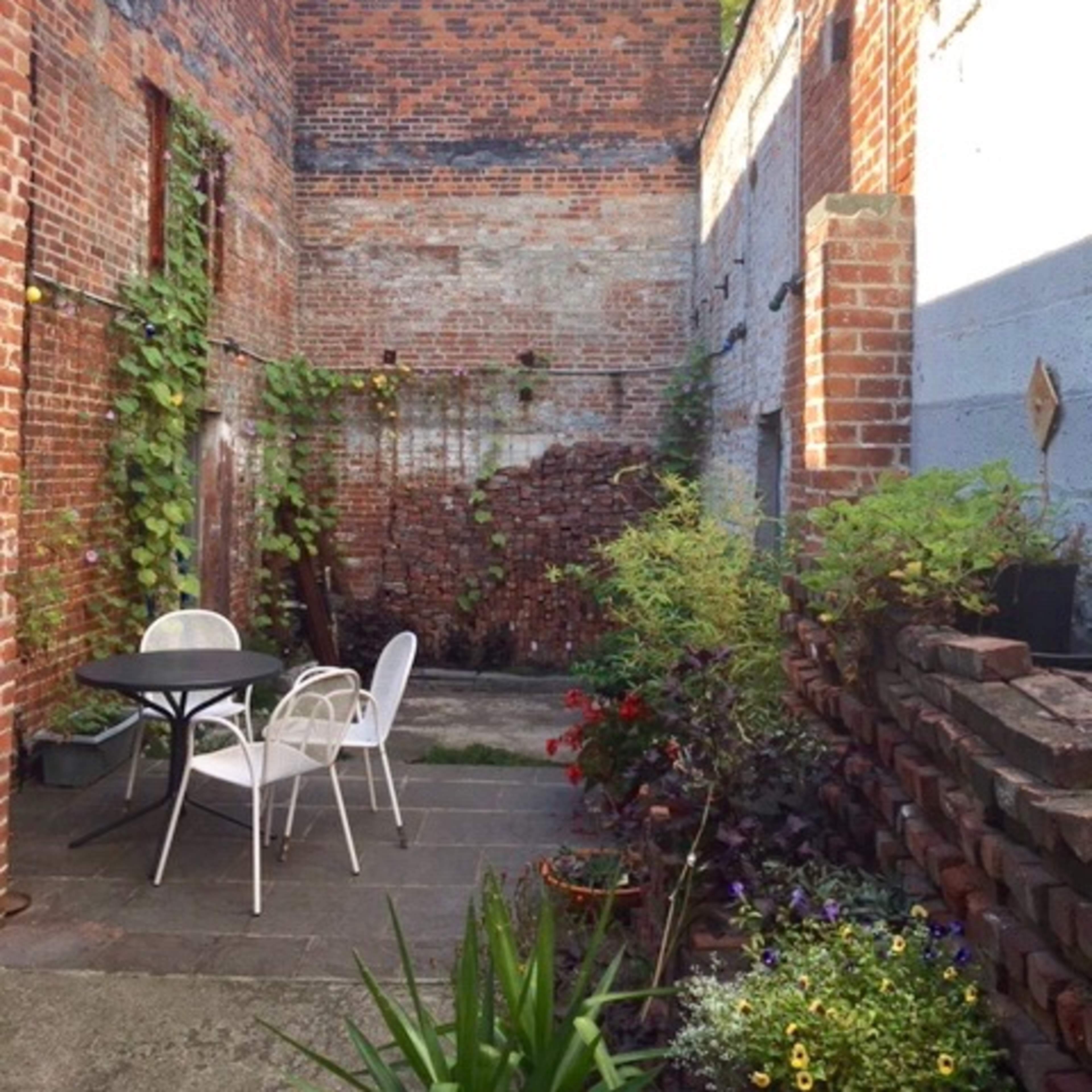 A small, brick-walled courtyard filled with greenery and a table set with white chairs.