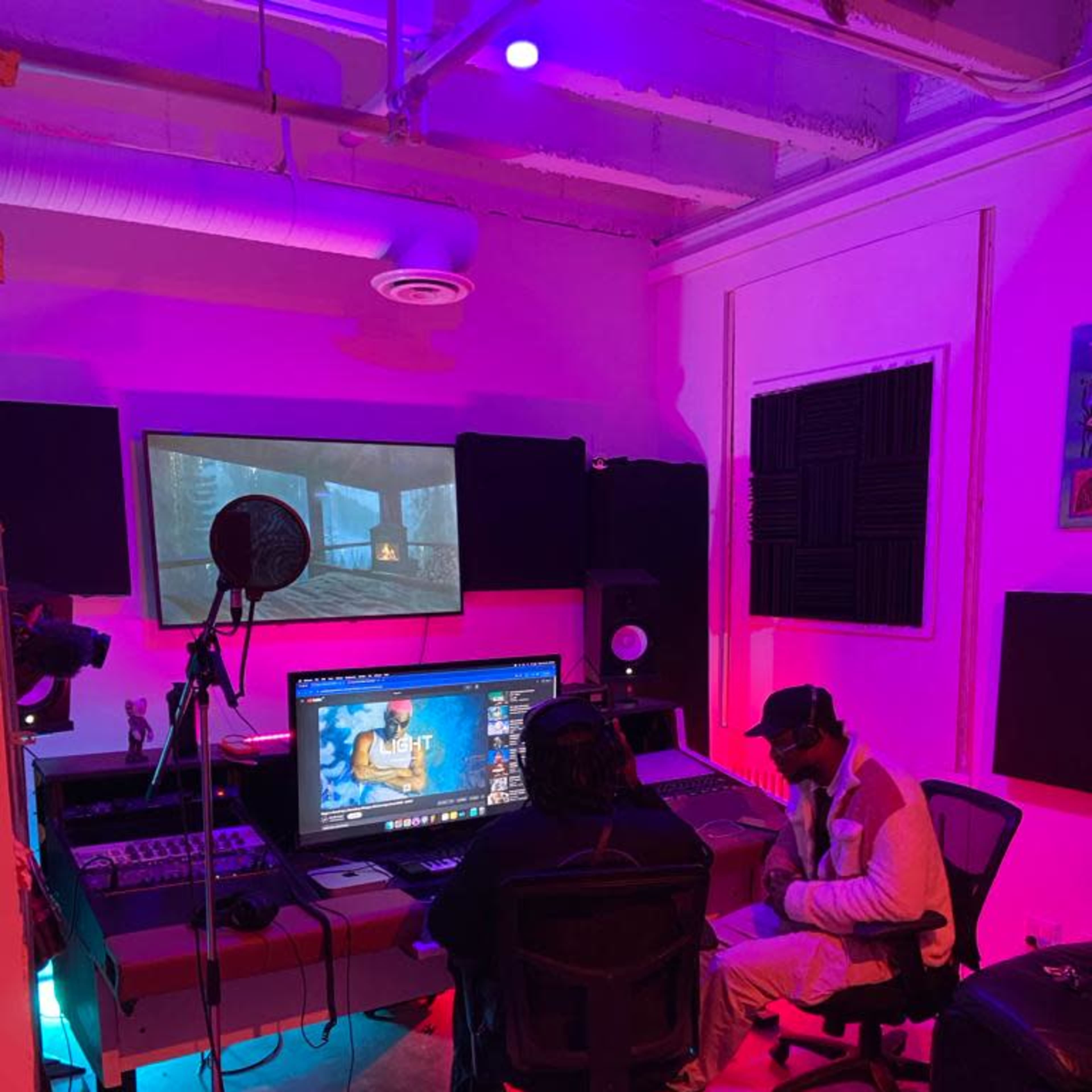 A music production studio features a computer setup with two people seated in front of multiple screens and audio equipment, illuminated by colorful lights.