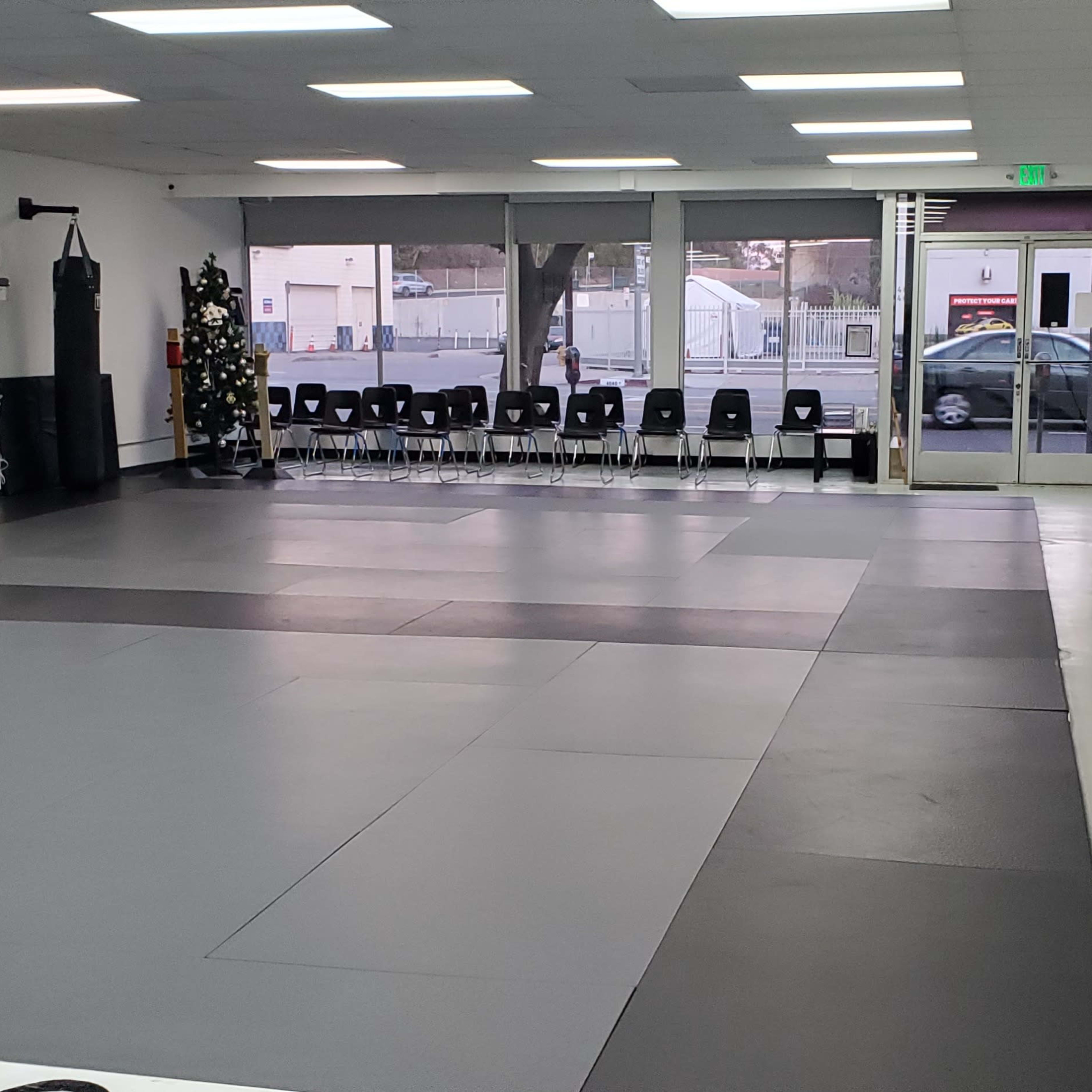 A spacious martial arts studio with gray mats, empty chairs along one wall, and large windows revealing an outside view.