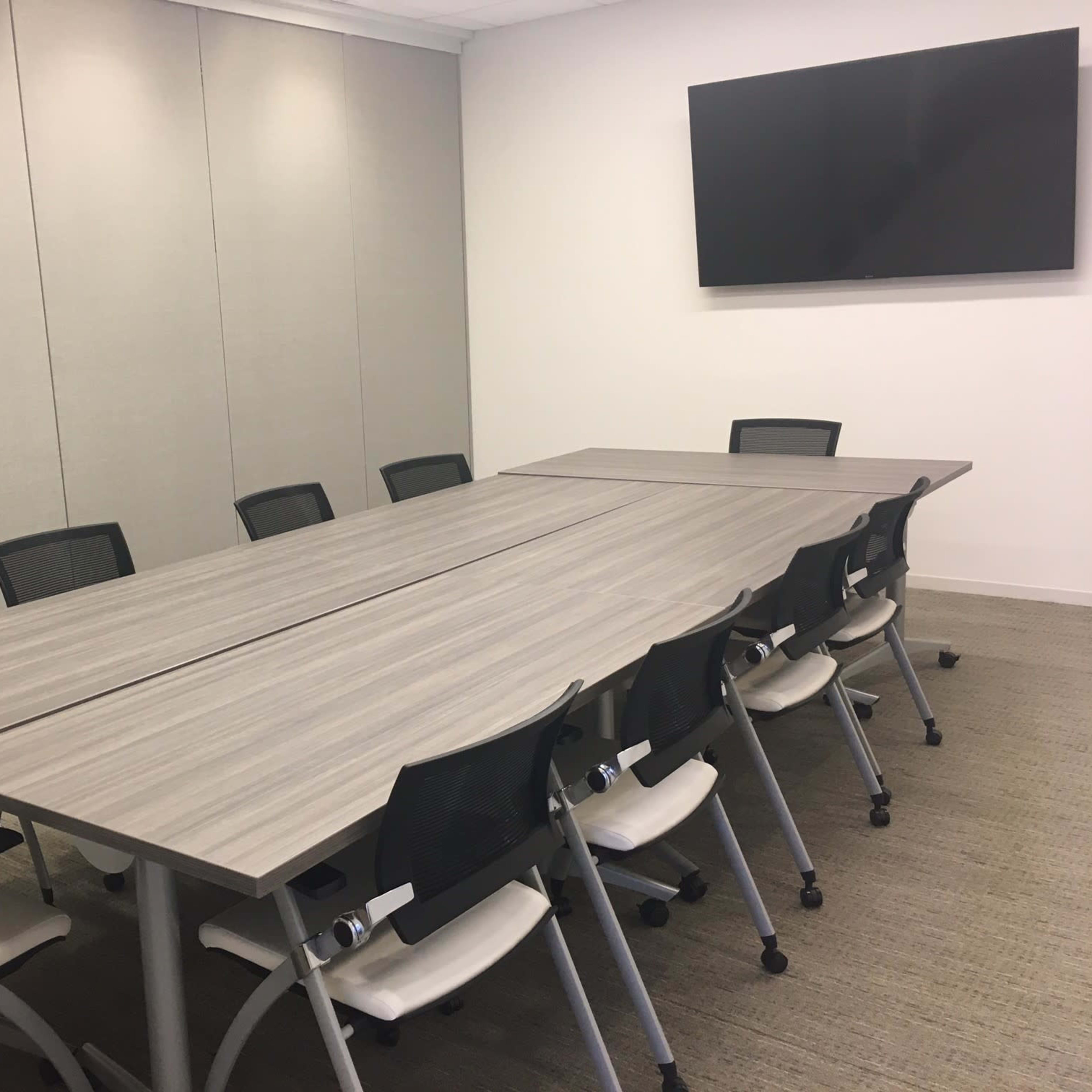 A large conference room features a long rectangular table with ten ergonomic chairs and a wall-mounted flat-screen television.