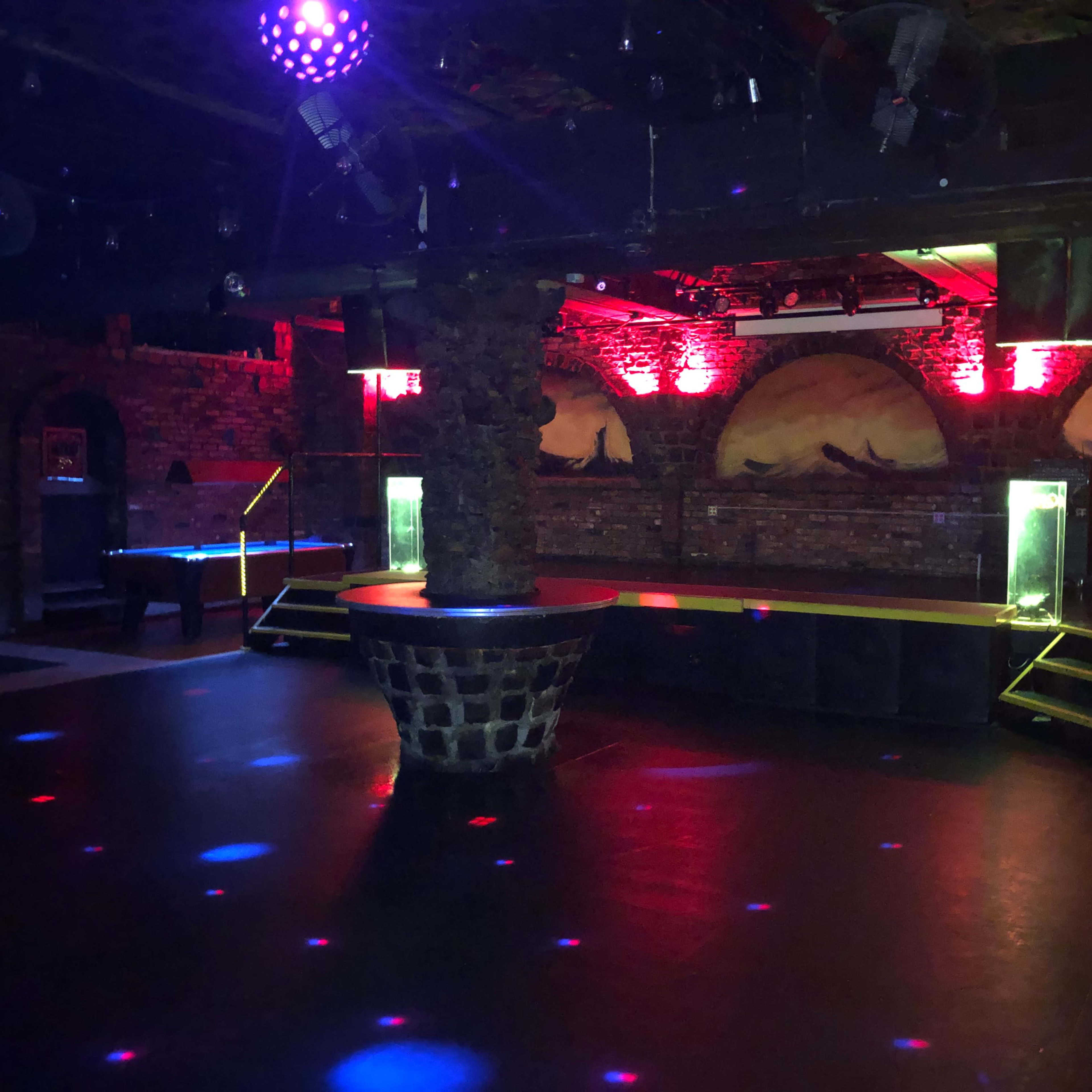 A dimly lit nightclub interior with a circular bar in the center, surrounded by illuminated stages and stone walls.