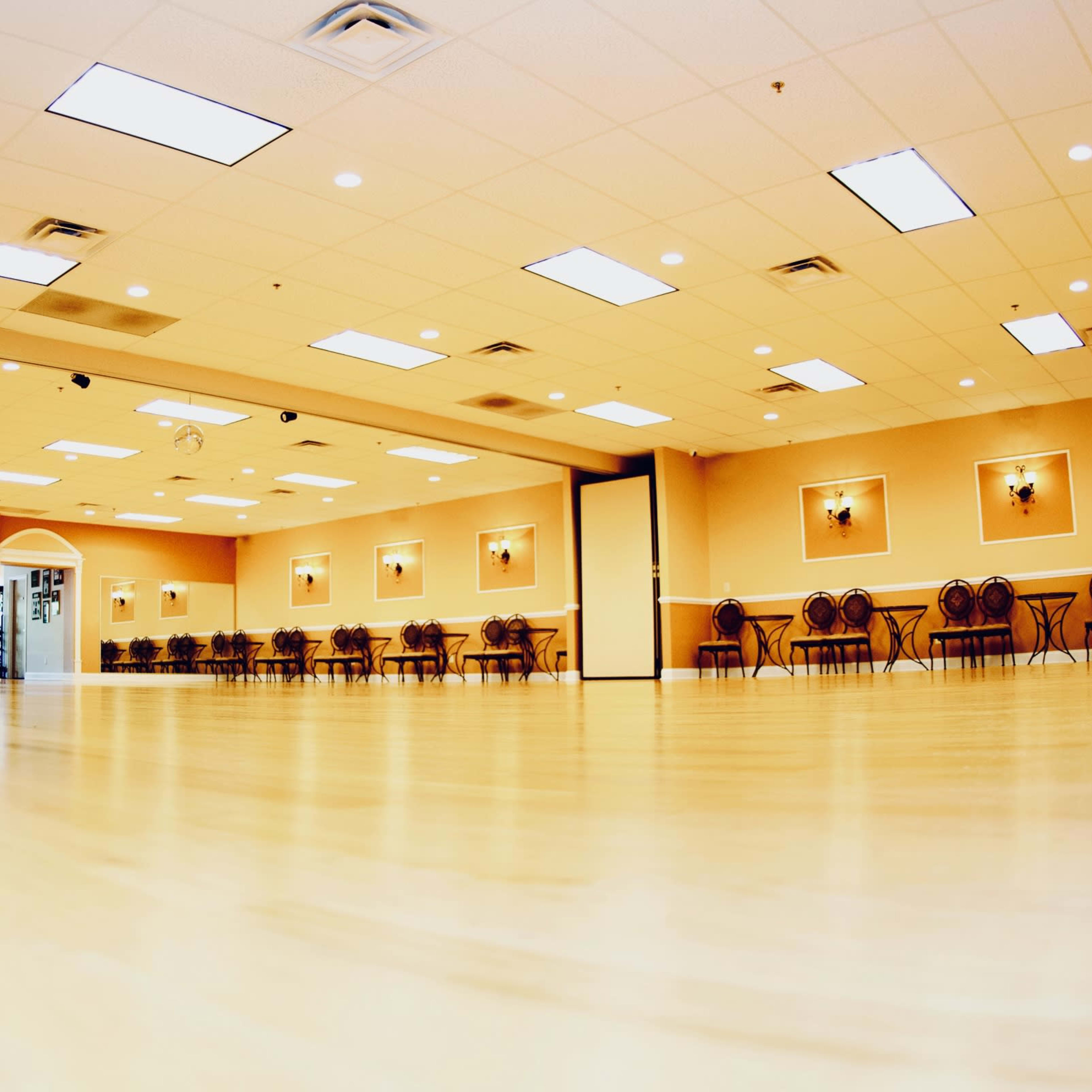 A spacious hall with wooden flooring, illuminated by overhead lights, and lined with chairs along the walls.