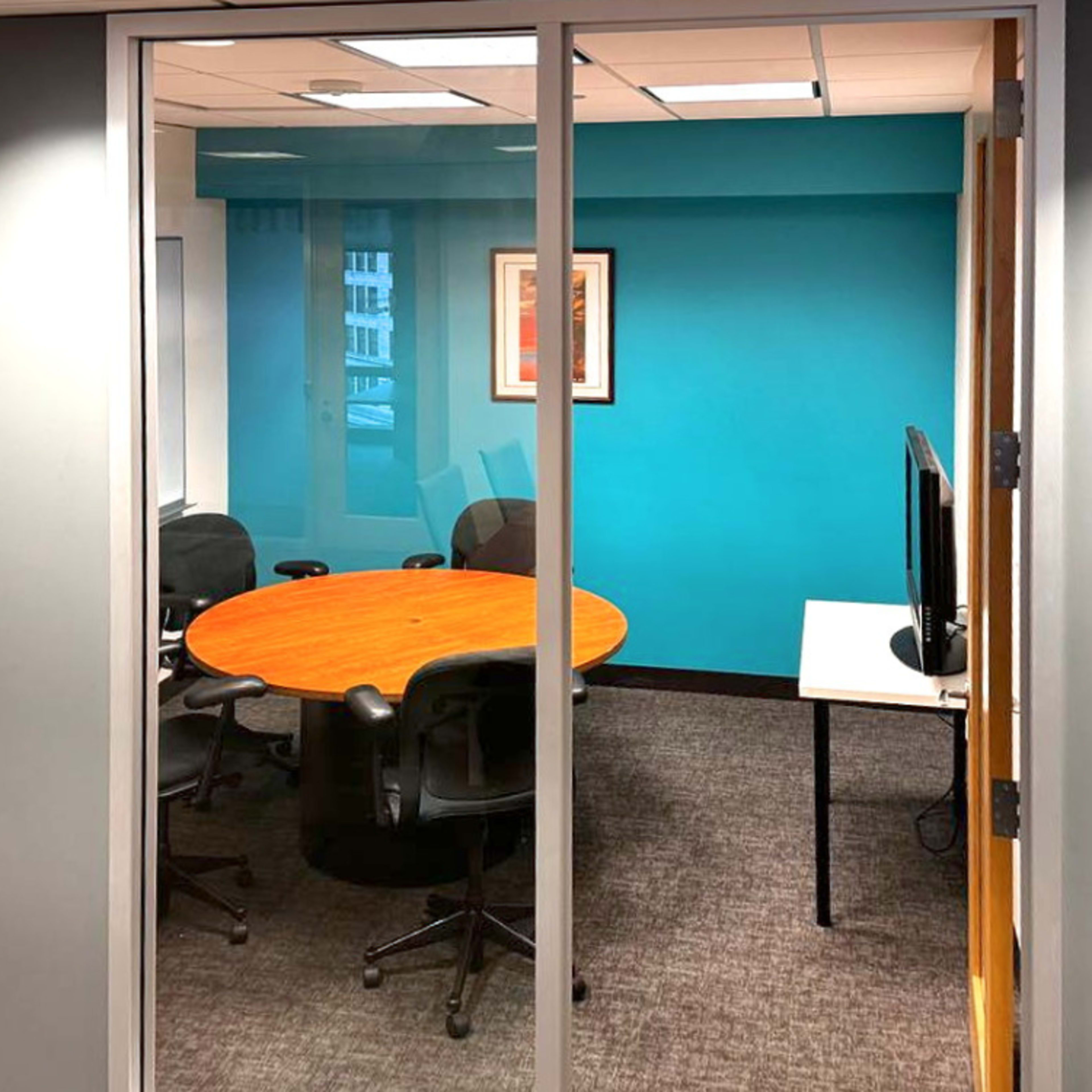 A glass-walled conference room with a round wooden table and rolling chairs, set against a turquoise wall.