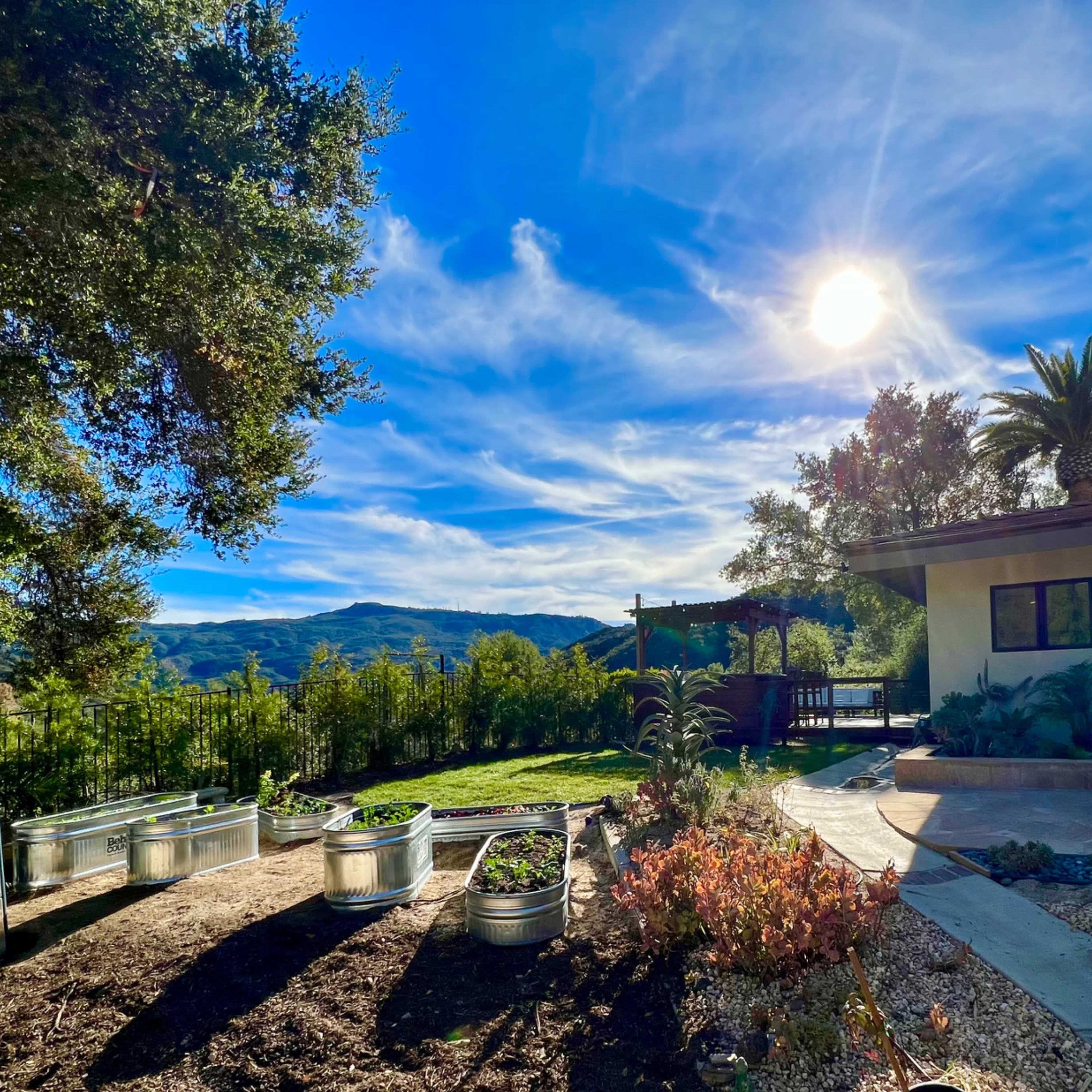 A garden area with metal planters, a greenhouse, and a house under a bright sun against a backdrop of blue skies and mountains.