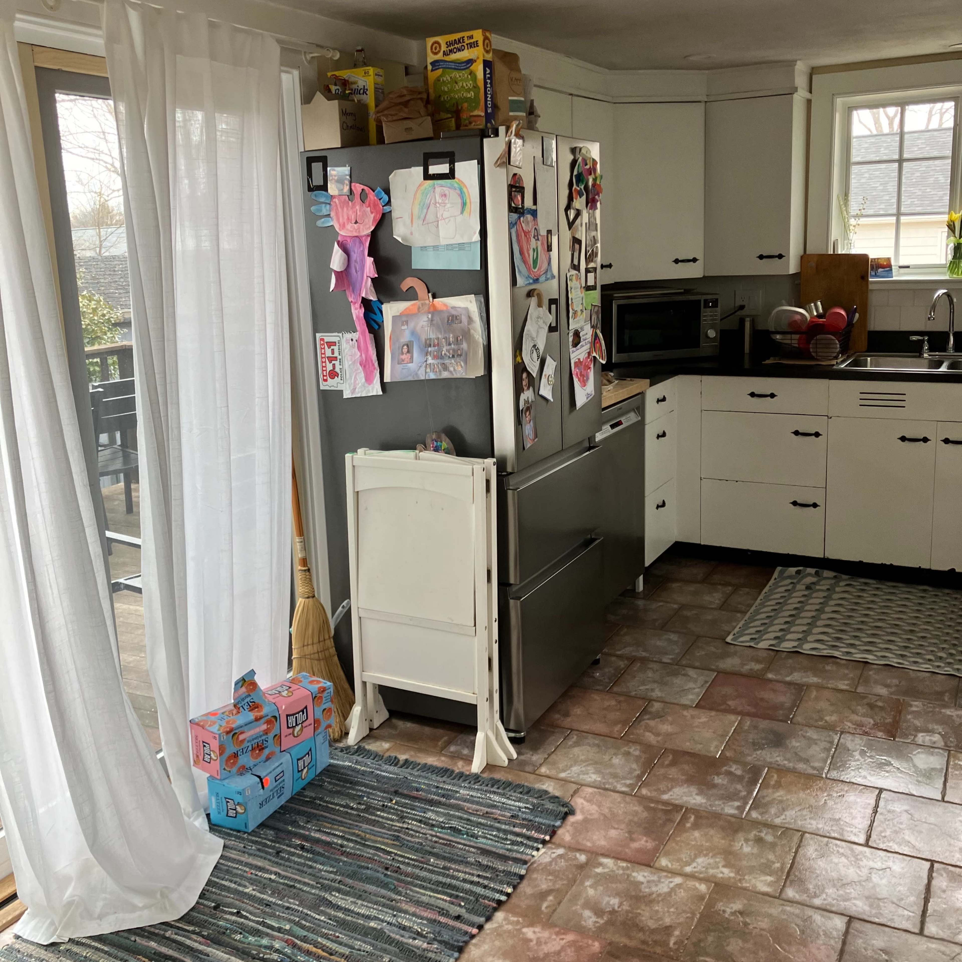 A kitchen with a refrigerator adorned with drawings, a microwave, and a door leading to an outdoor area, all set on tiled flooring.