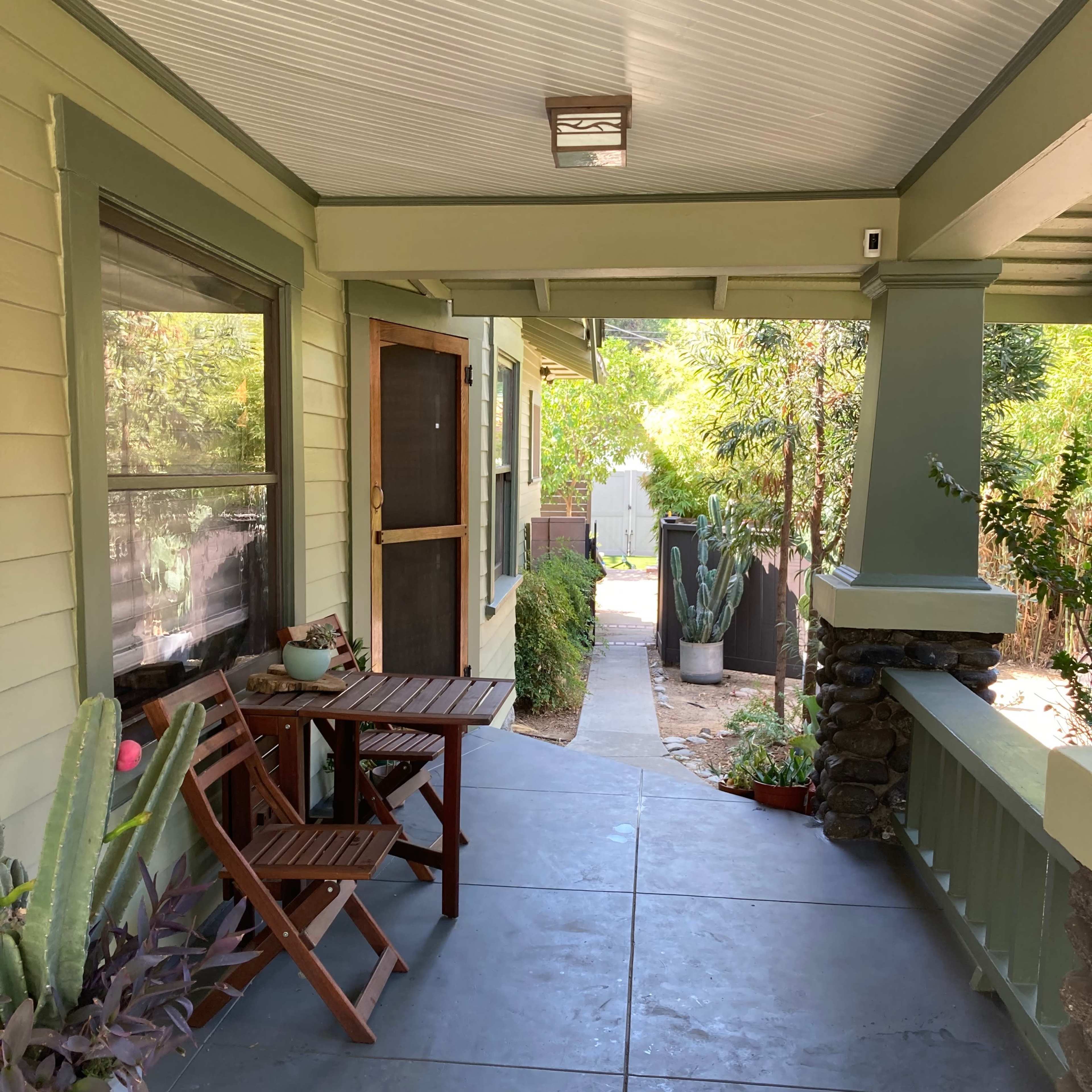 A porch with a wooden folding table and chairs, surrounded by potted plants and a pathway leading to a garden.
