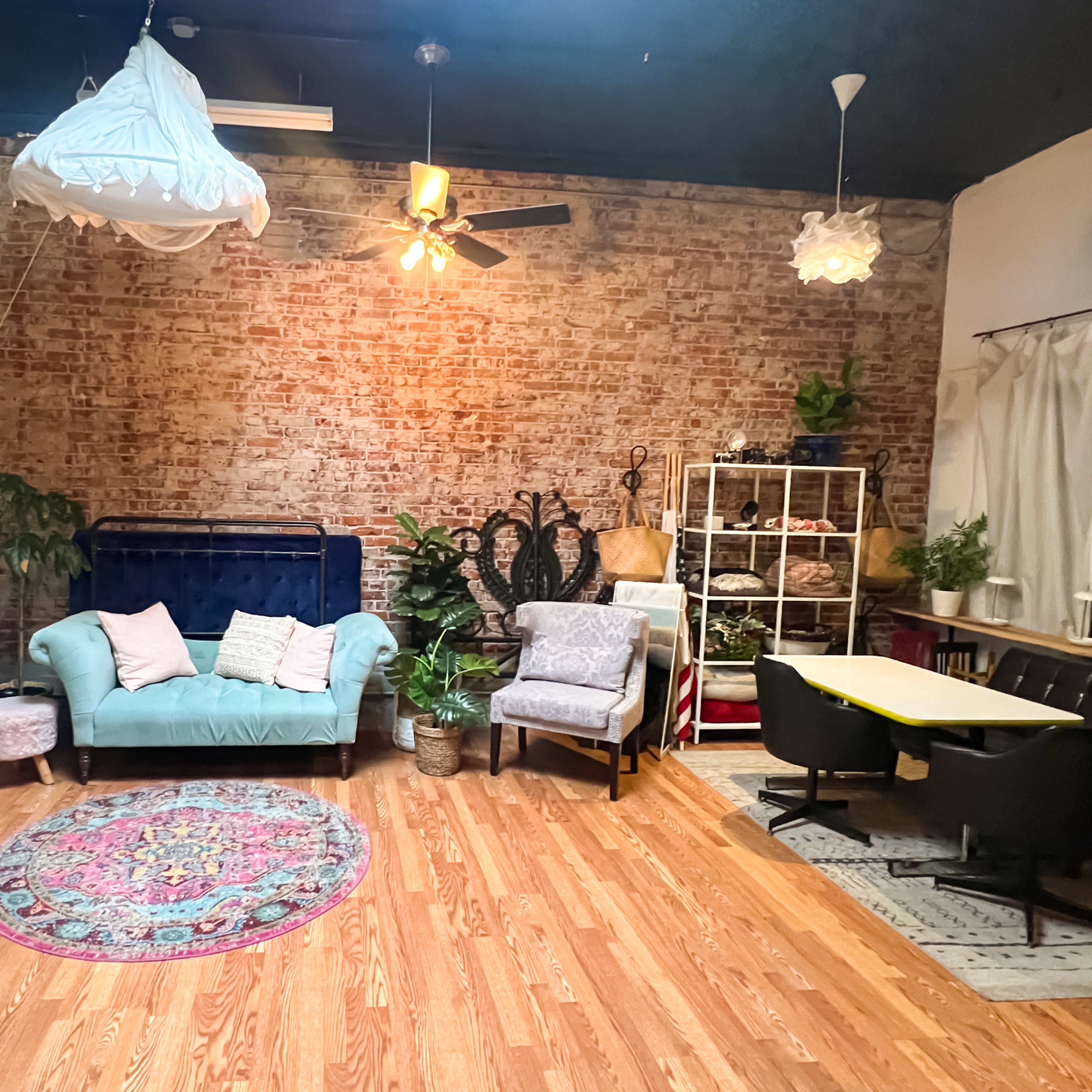 A spacious room with exposed brick walls, featuring a mix of seating arrangements, houseplants, and decorative rugs.