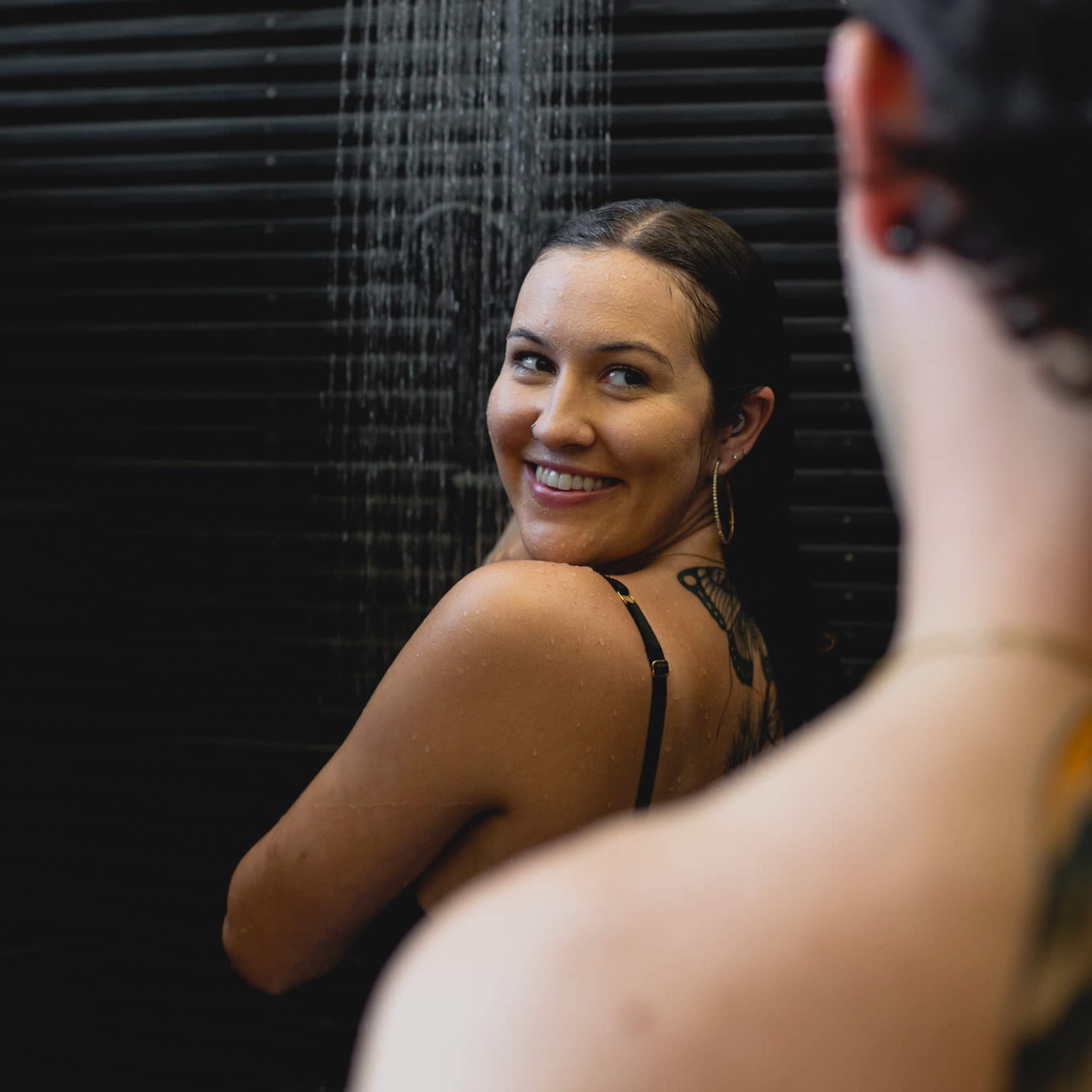 A woman with wet hair smiles while standing under a shower, as a person partially visible in the foreground looks on.