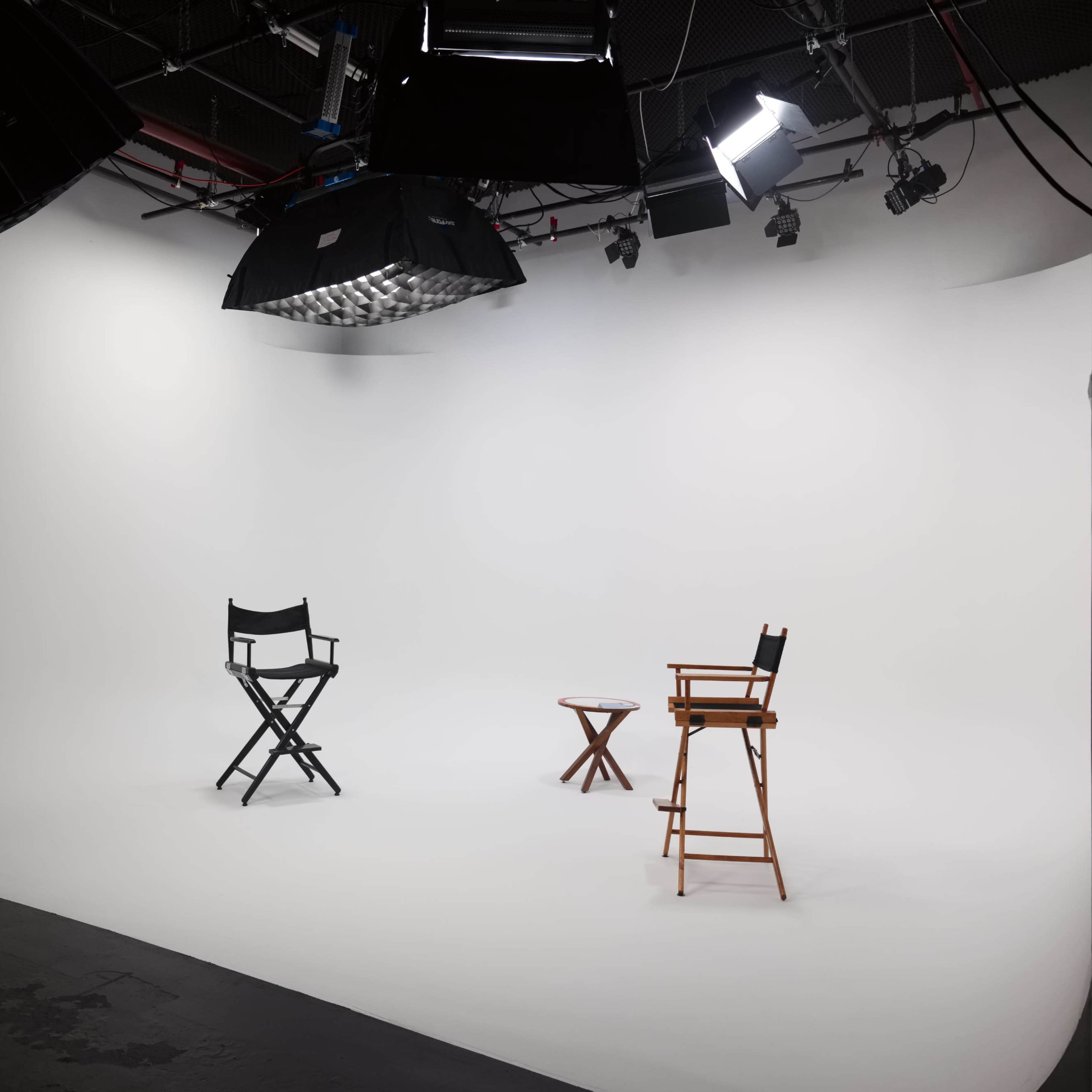 A photography studio with a seamless white backdrop, two director's chairs, and a small table placed on a dark floor.