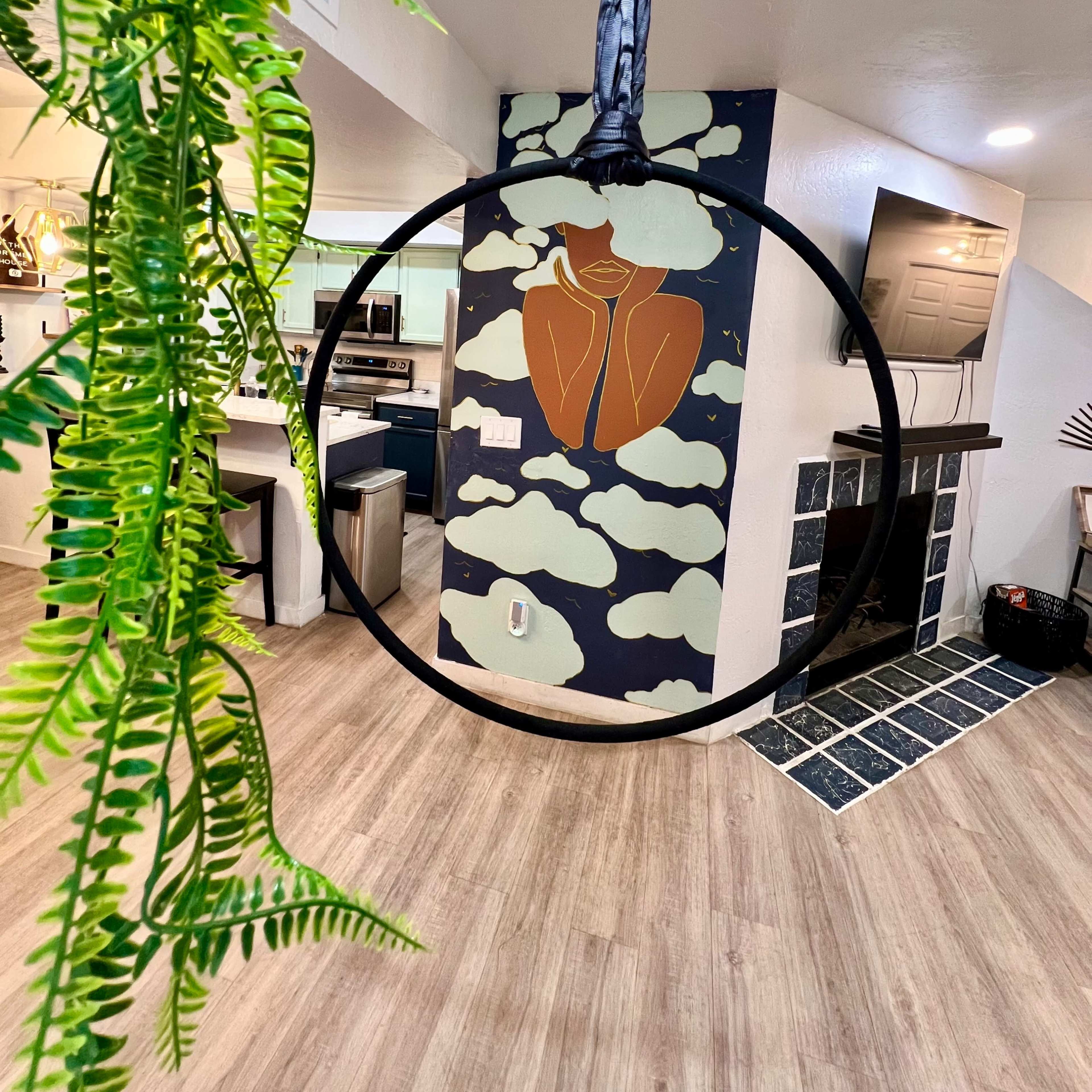 A circular hanging decor piece in a room with a mural of a figure surrounded by clouds on the wall and a plant in the foreground.