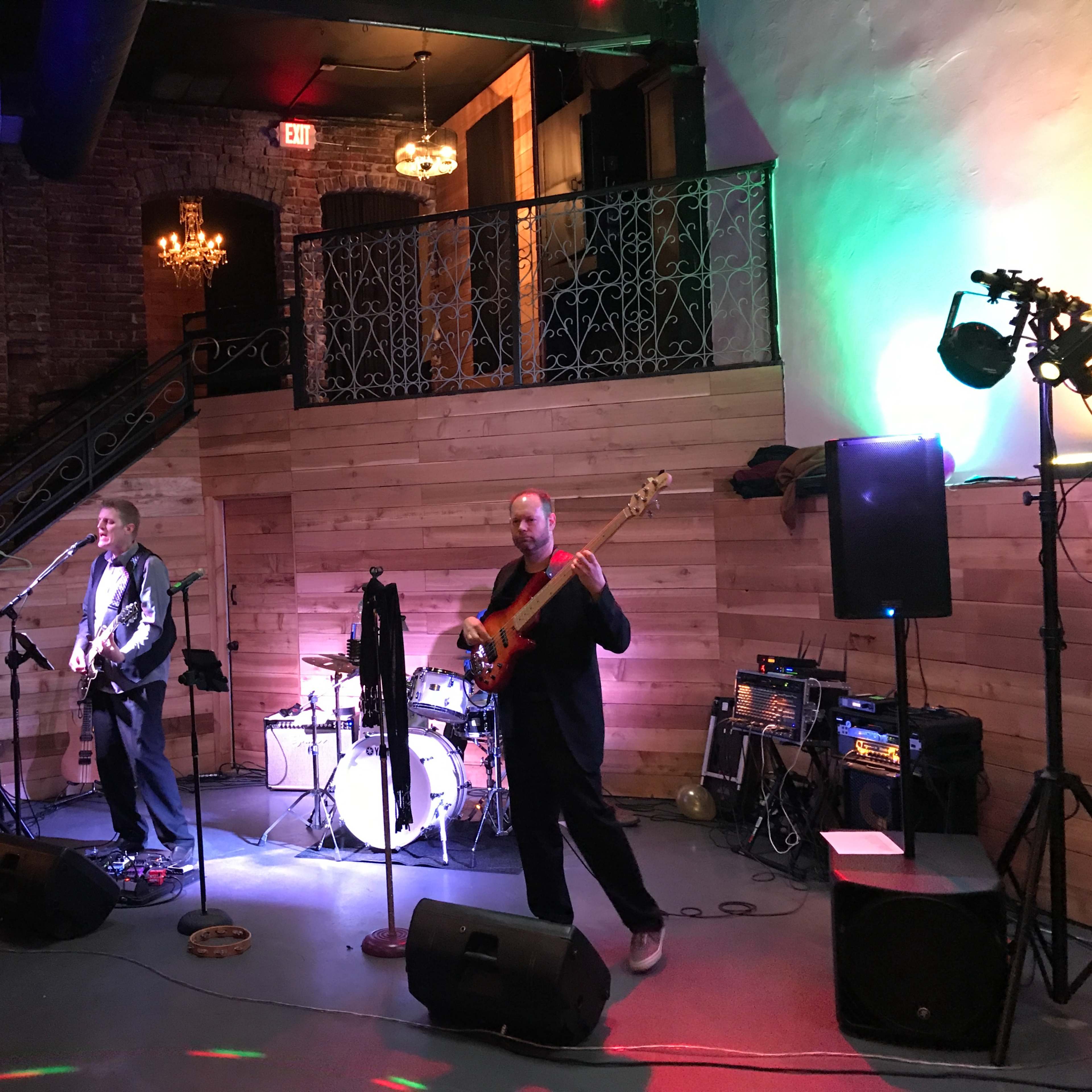 A band performs on stage in a venue with brick walls and dim lighting, featuring instruments like a guitar, bass, and drums.