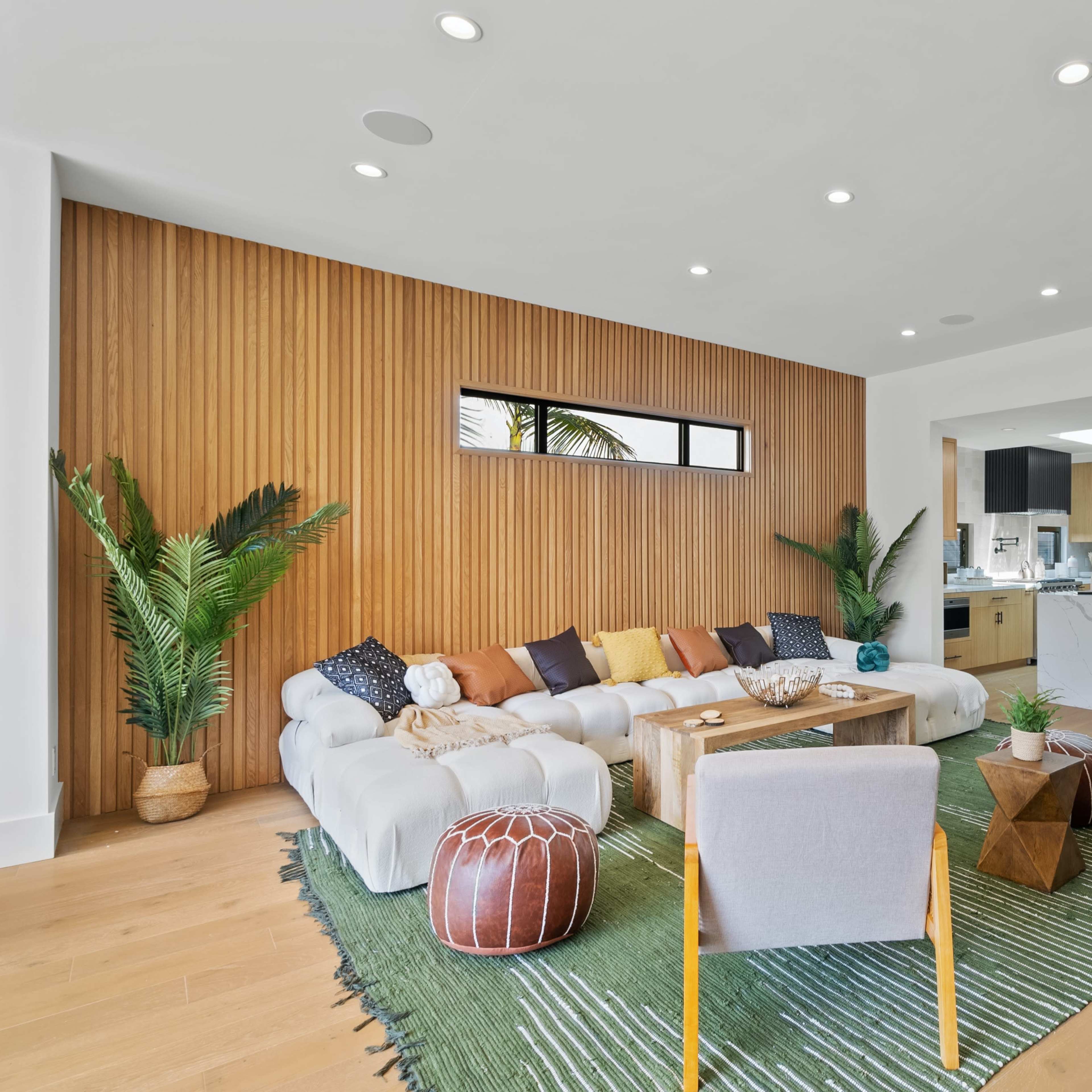 A modern living room with a sectional sofa, a wooden accent wall, a coffee table, and greenery, leading to an open kitchen and dining area.