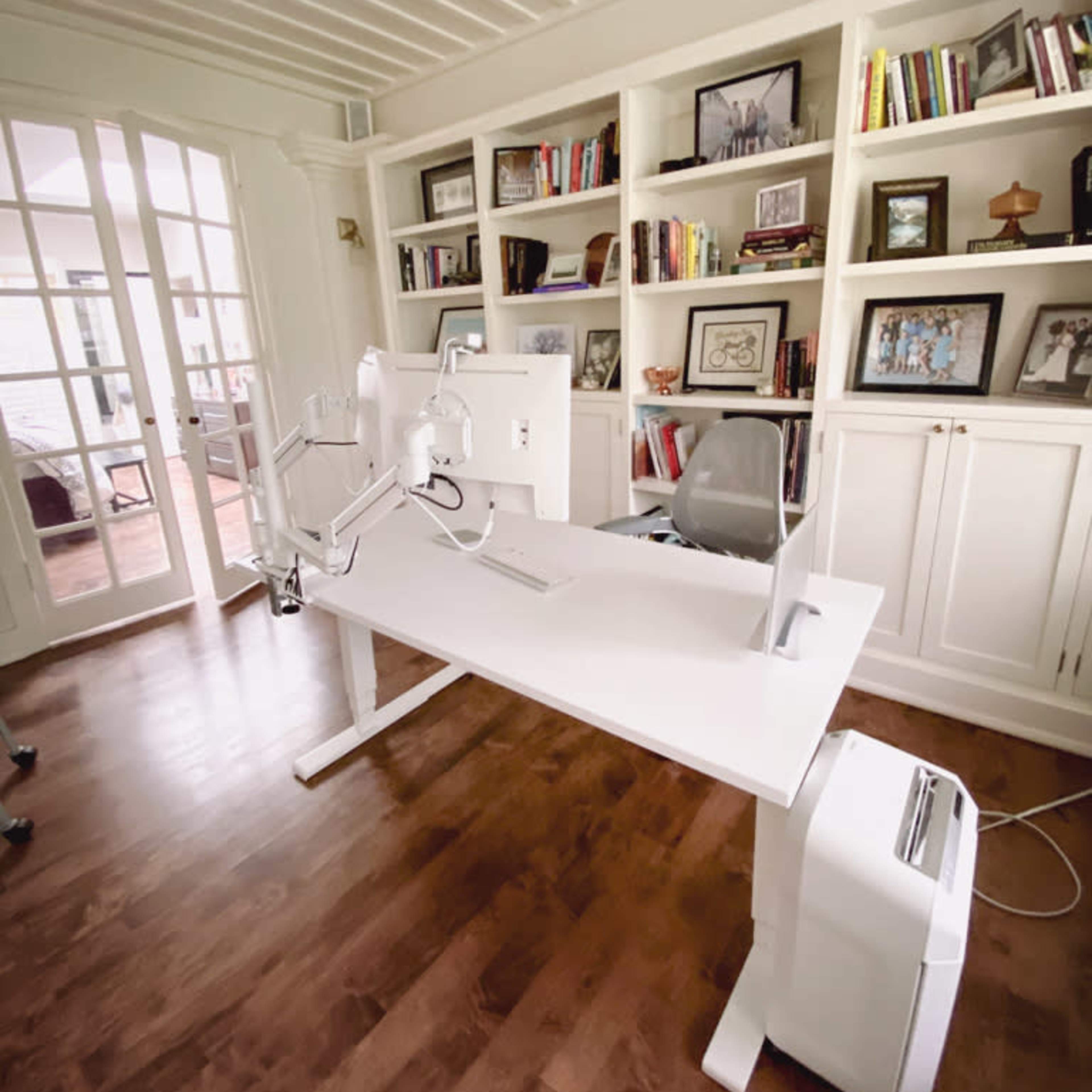 A modern white desk with a dual-monitor setup is positioned in a well-lit room featuring built-in bookshelves filled with framed photos and books.