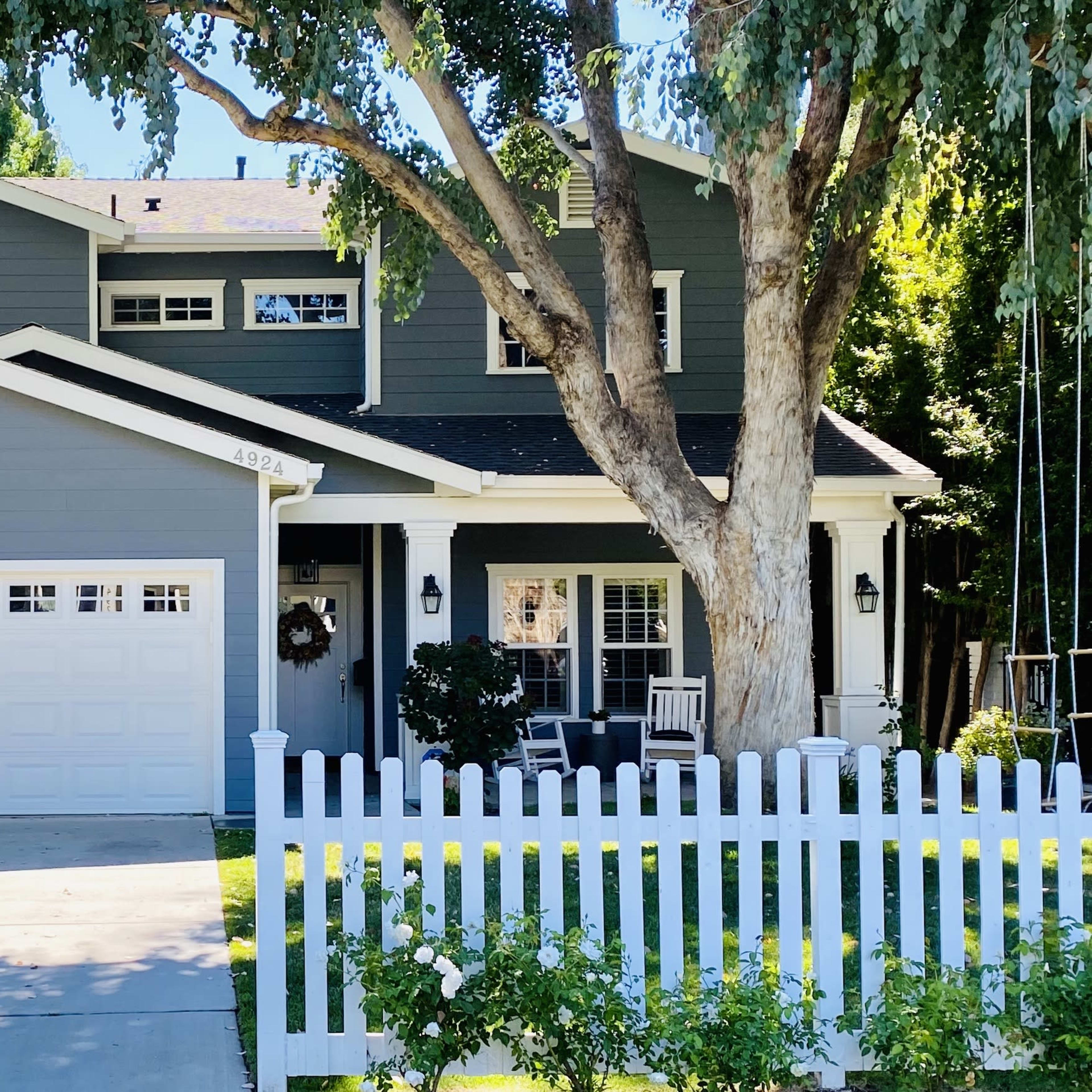 A two-story gray house with a white picket fence, a concrete driveway, and a large tree in the front yard.