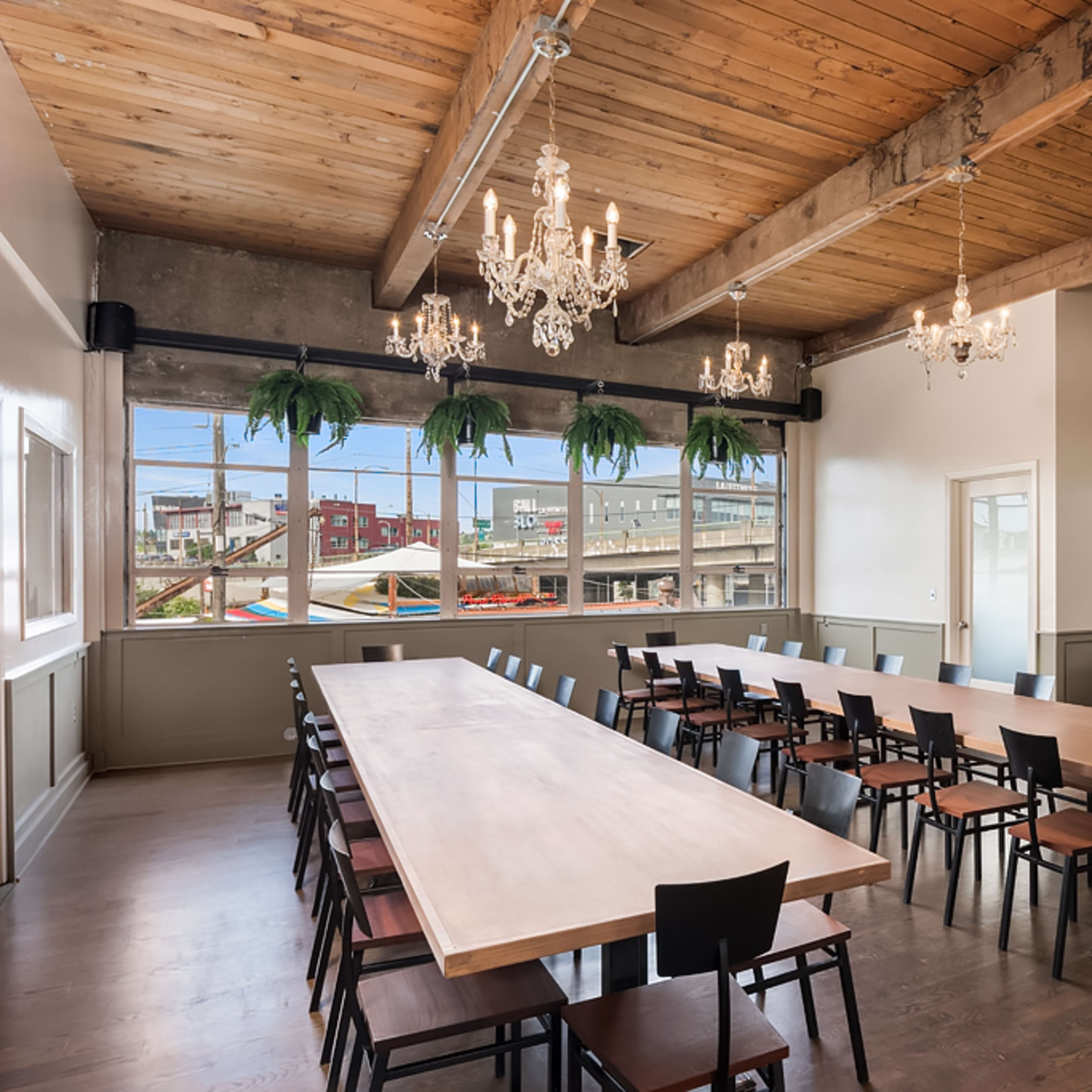 A conference room features a long wooden table surrounded by black chairs, with chandeliers hanging from the ceiling and large windows displaying an outdoor view.
