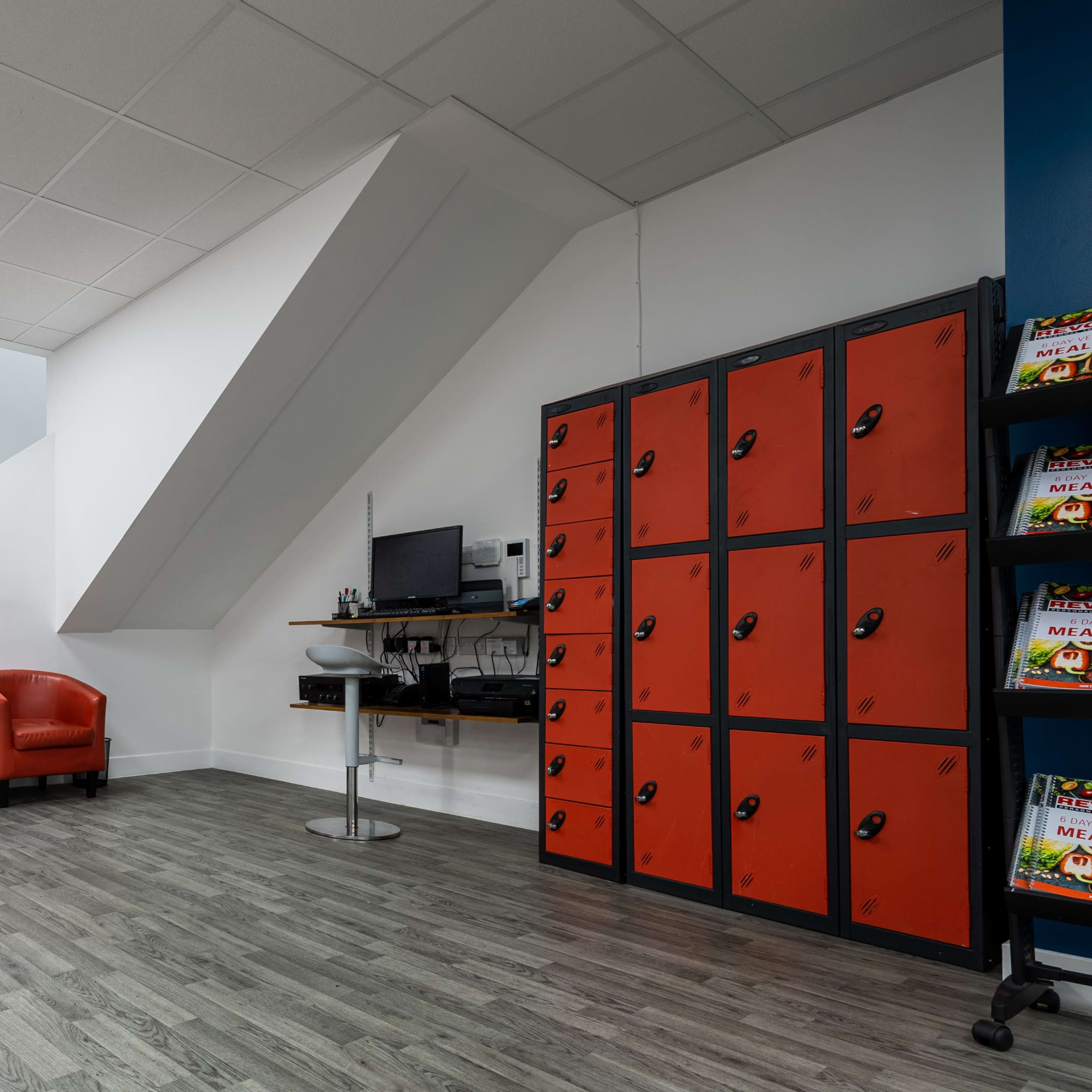 A room with two red chairs separated by a small table, a staircase on one side, orange lockers on the wall, and a wall-mounted monitor on a desk.