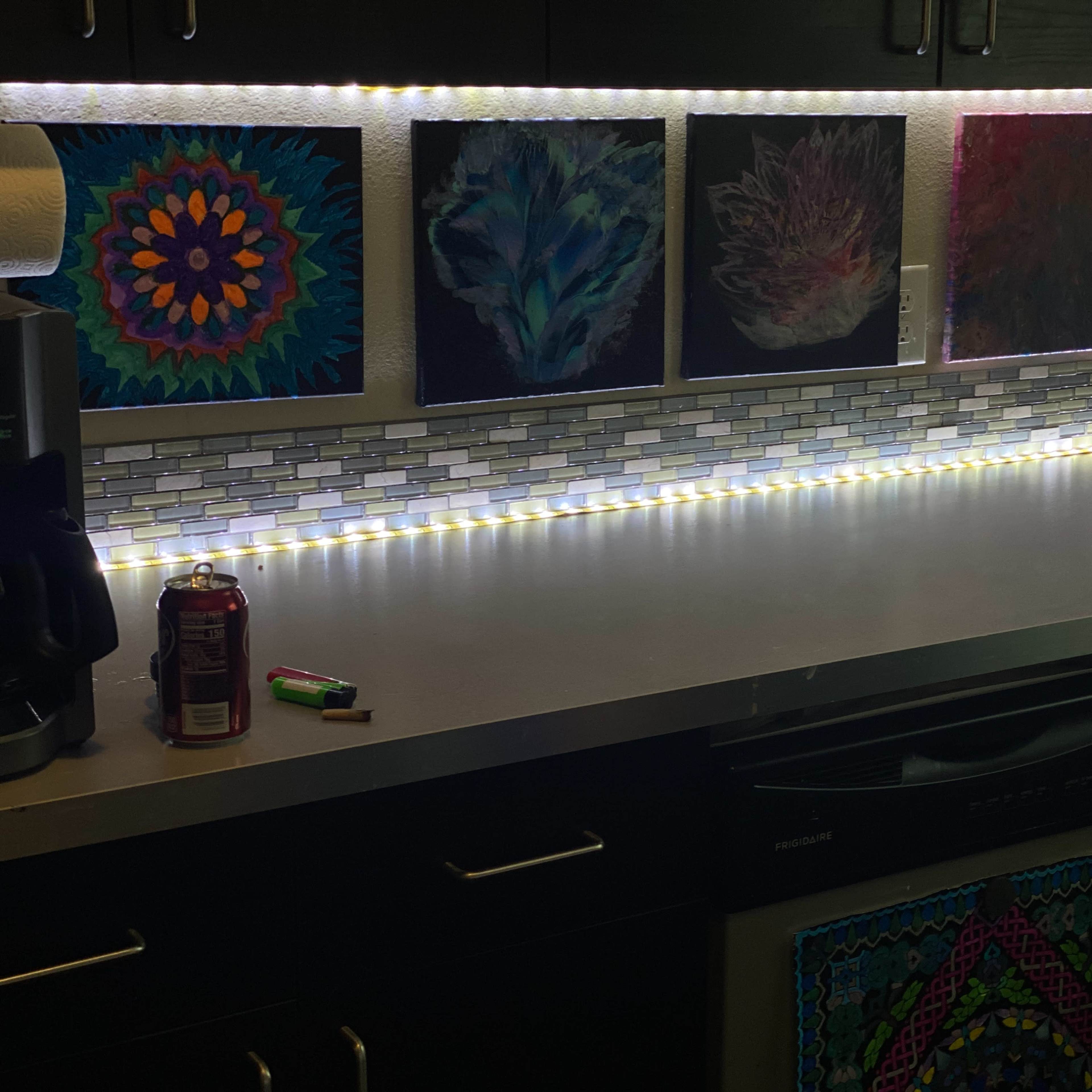 A kitchen counter with illuminated artwork above a backsplash, complemented by a coffee maker and a soft drink can on the surface.