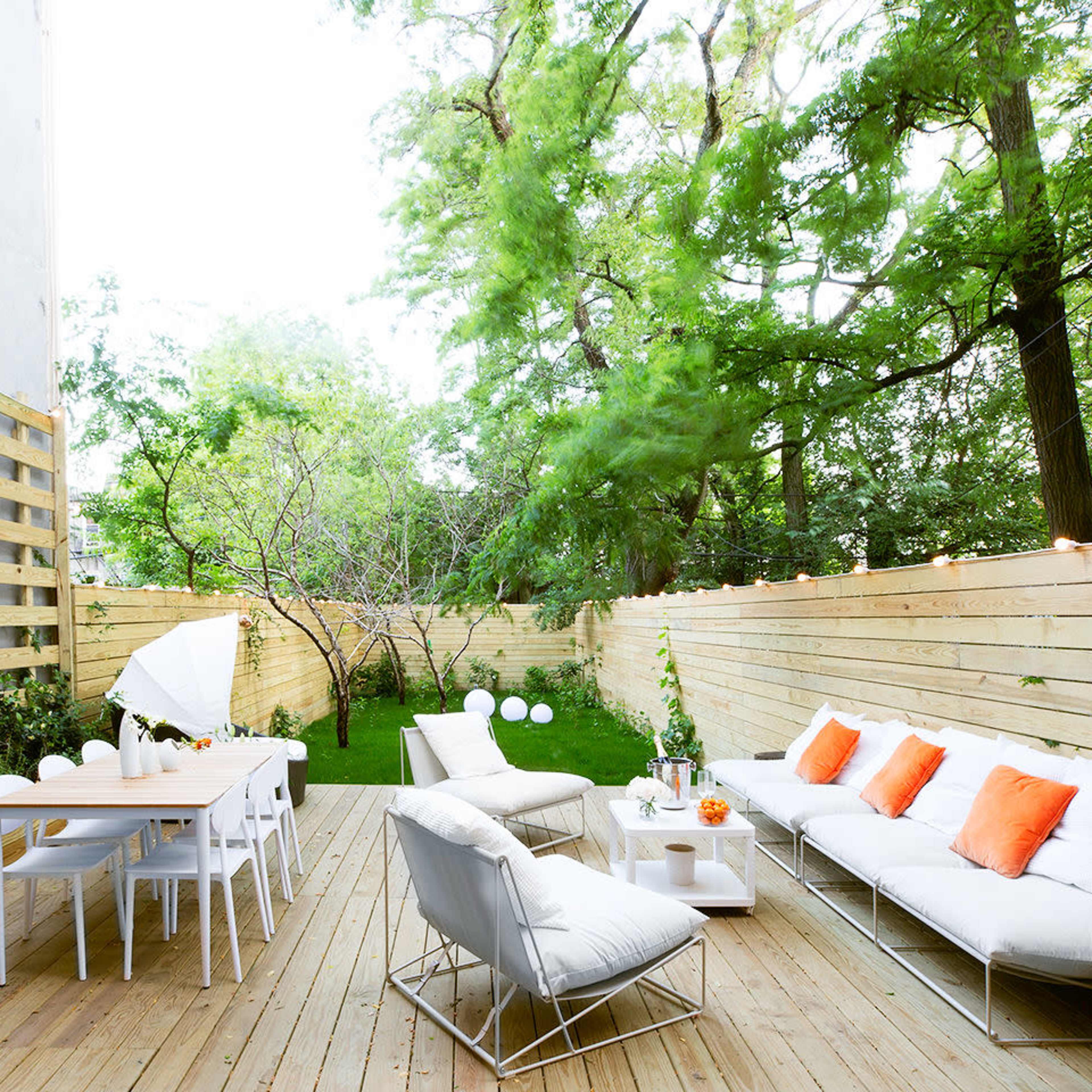 A wooden deck surrounded by greenery, featuring a seating area with white sofas and orange cushions, a dining table, and decorative plants.