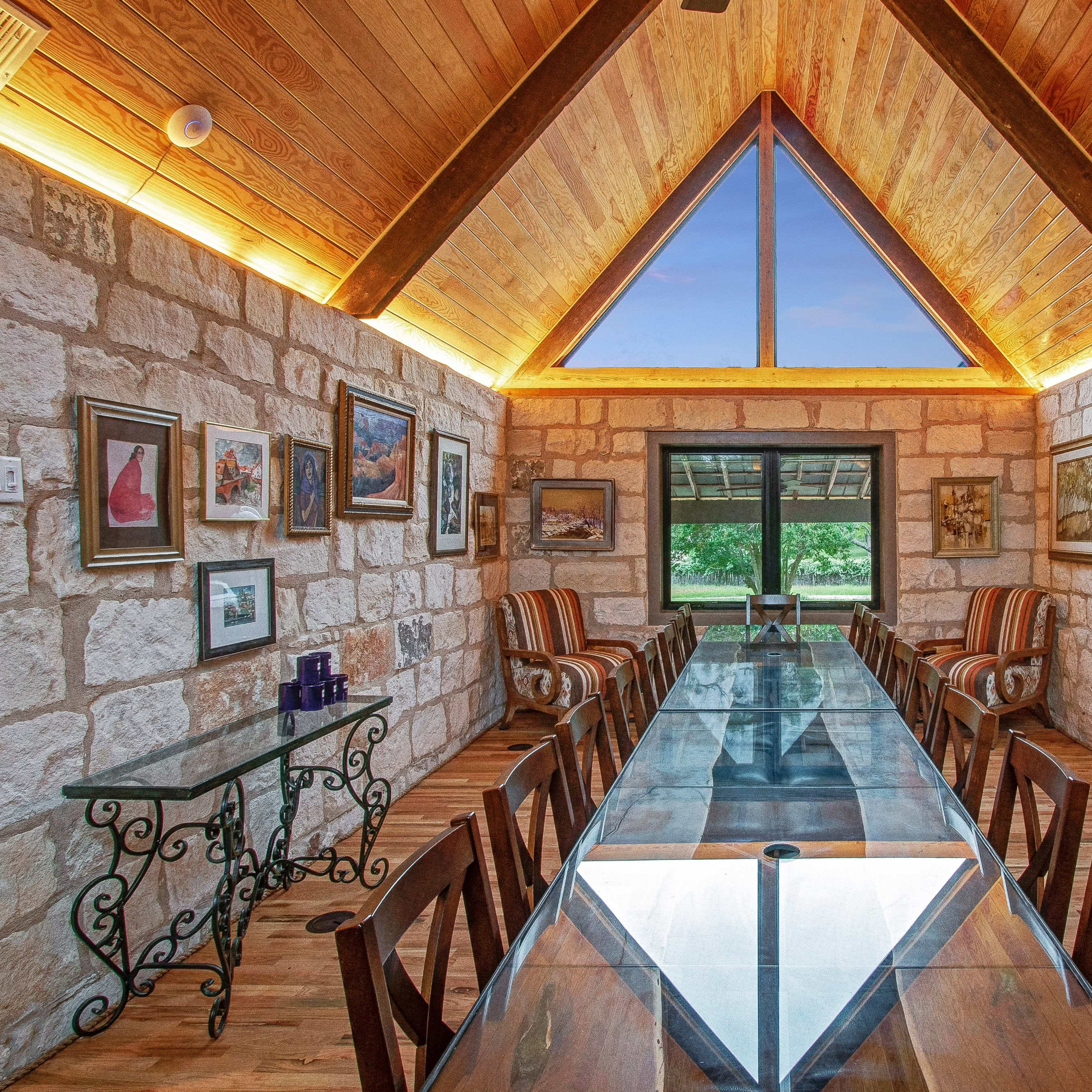 A modern dining room features a glass table surrounded by wooden chairs, stone walls, and large windows under a vaulted wooden ceiling.